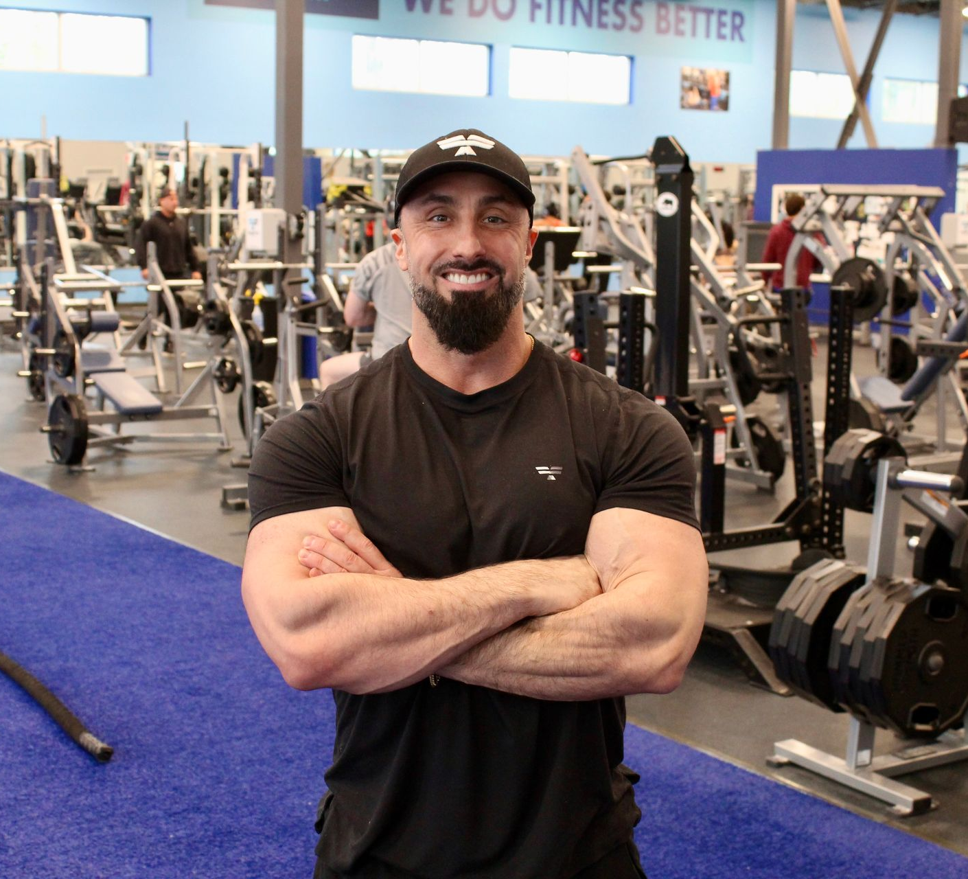 Man with arms crossed at a gym, wearing a black shirt and hat, surrounded by exercise equipment.