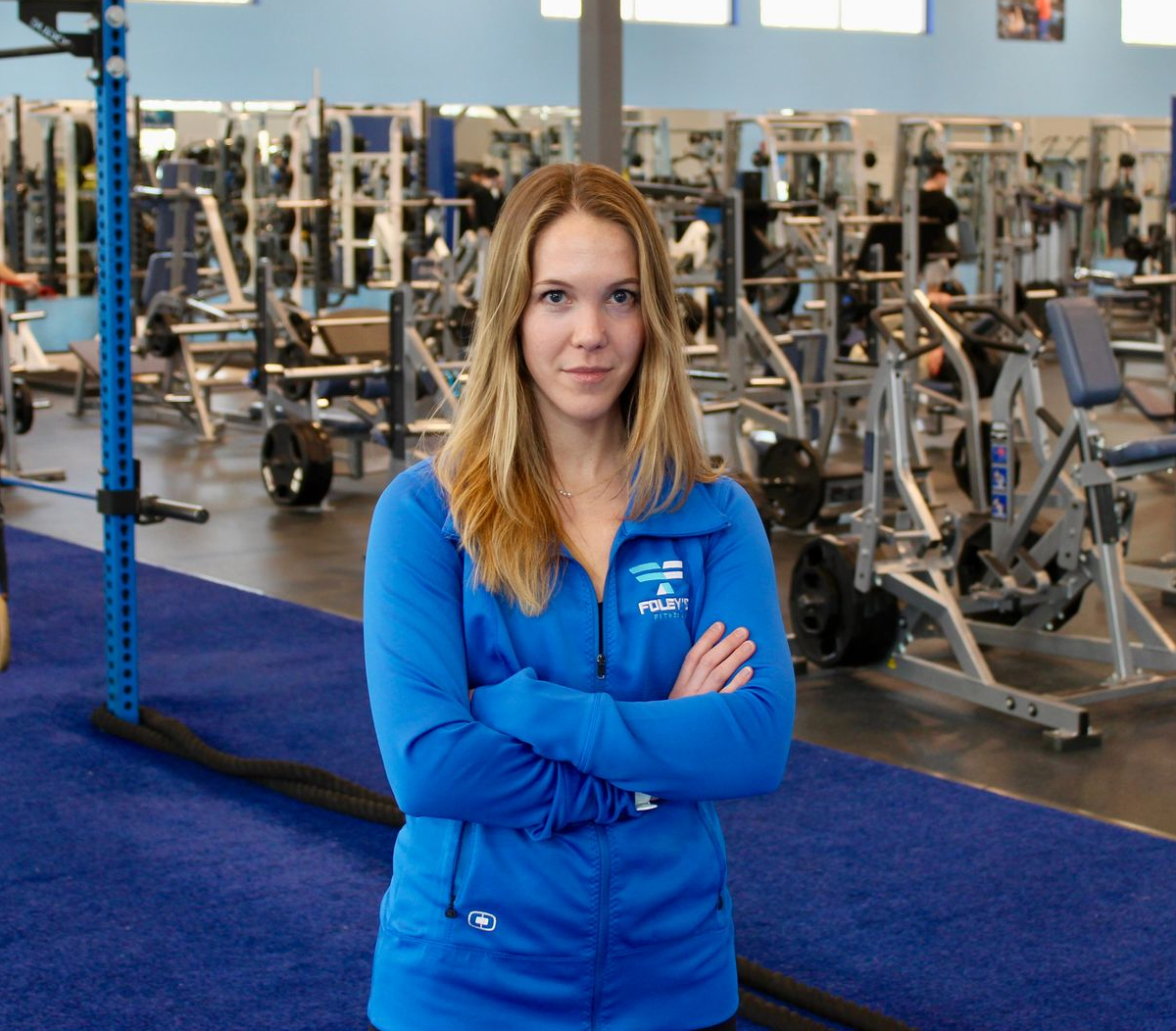 Woman in blue jacket with arms crossed stands in a gym, surrounded by equipment.