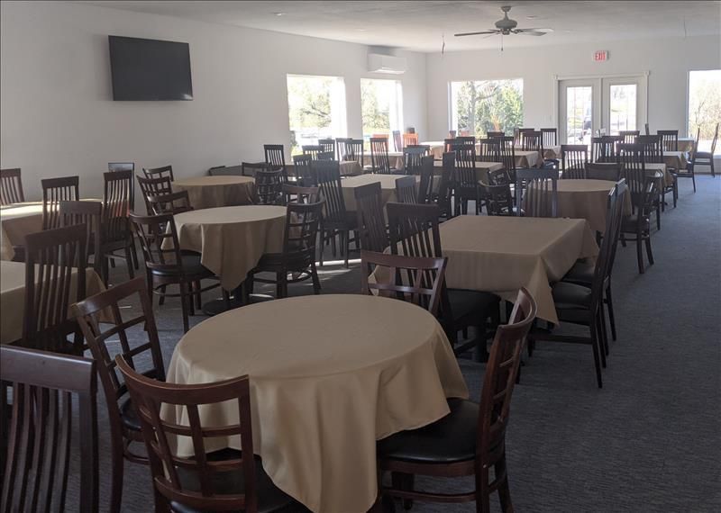 Empty dining hall with round tables, wooden chairs, and large windows letting in daylight