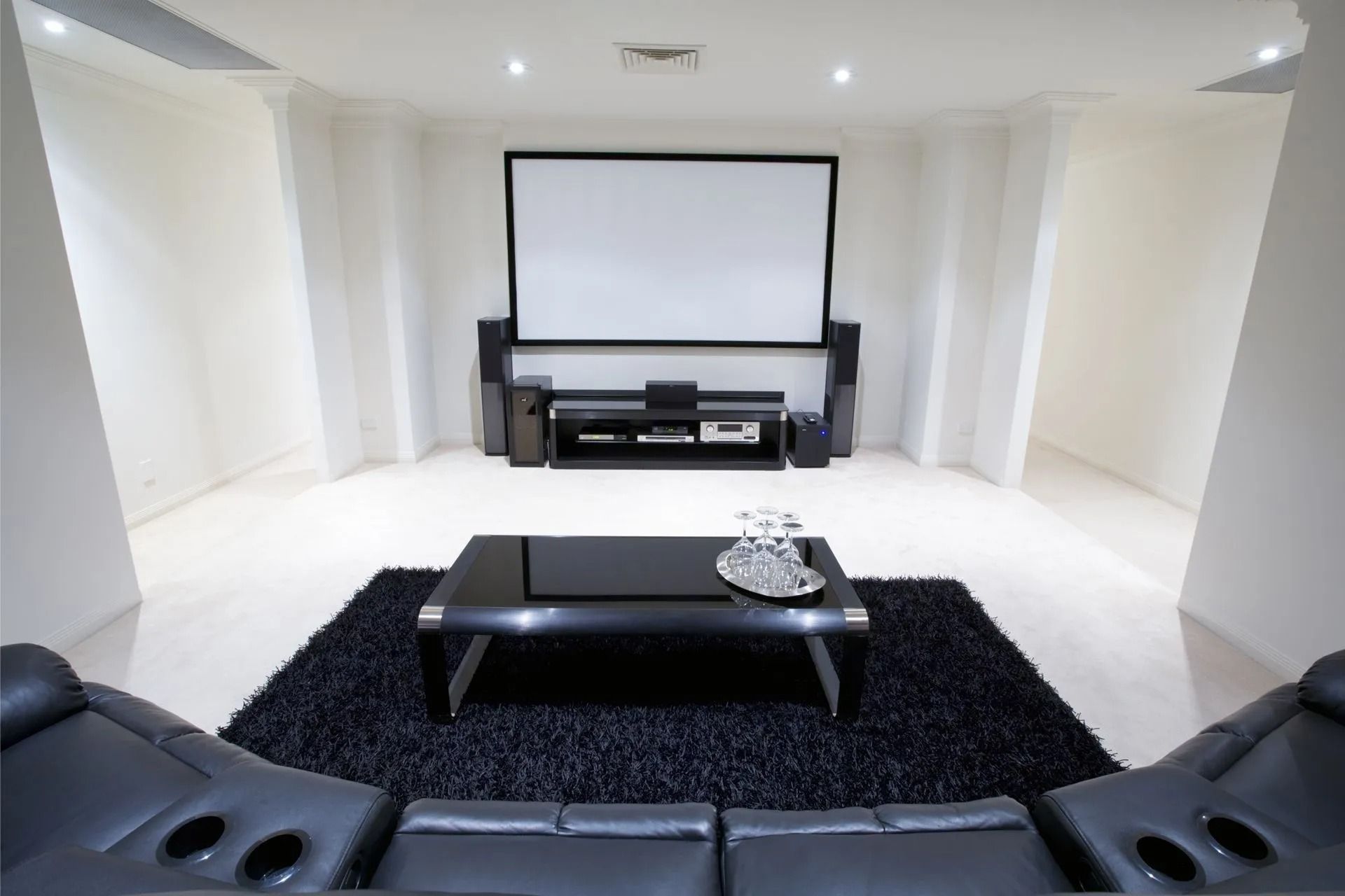 Modern home theater with white walls, large projection screen, black leather seating, and low coffee table