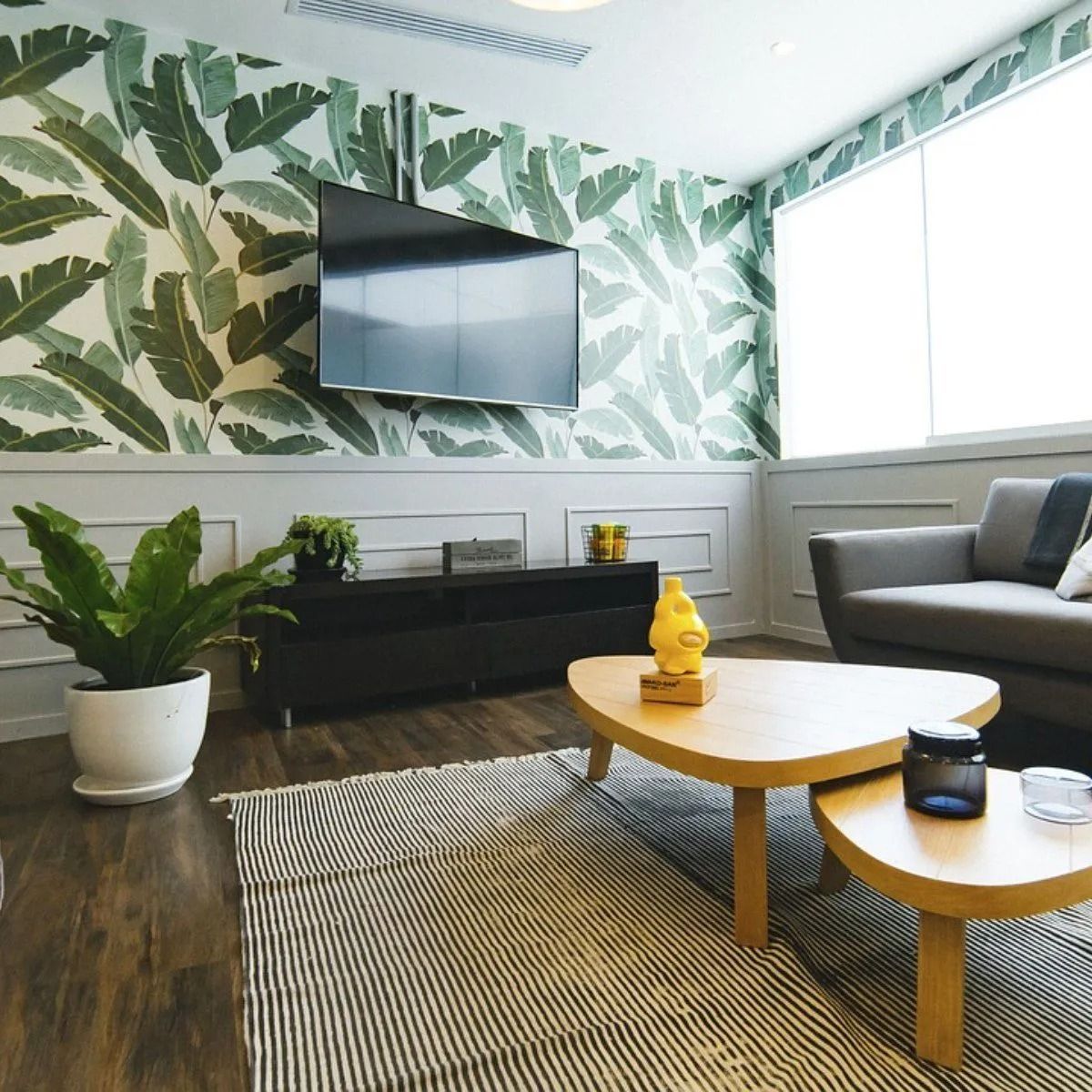 Bright living room with leaf wallpaper, wall TV, gray sofa, coffee tables, and potted plants.