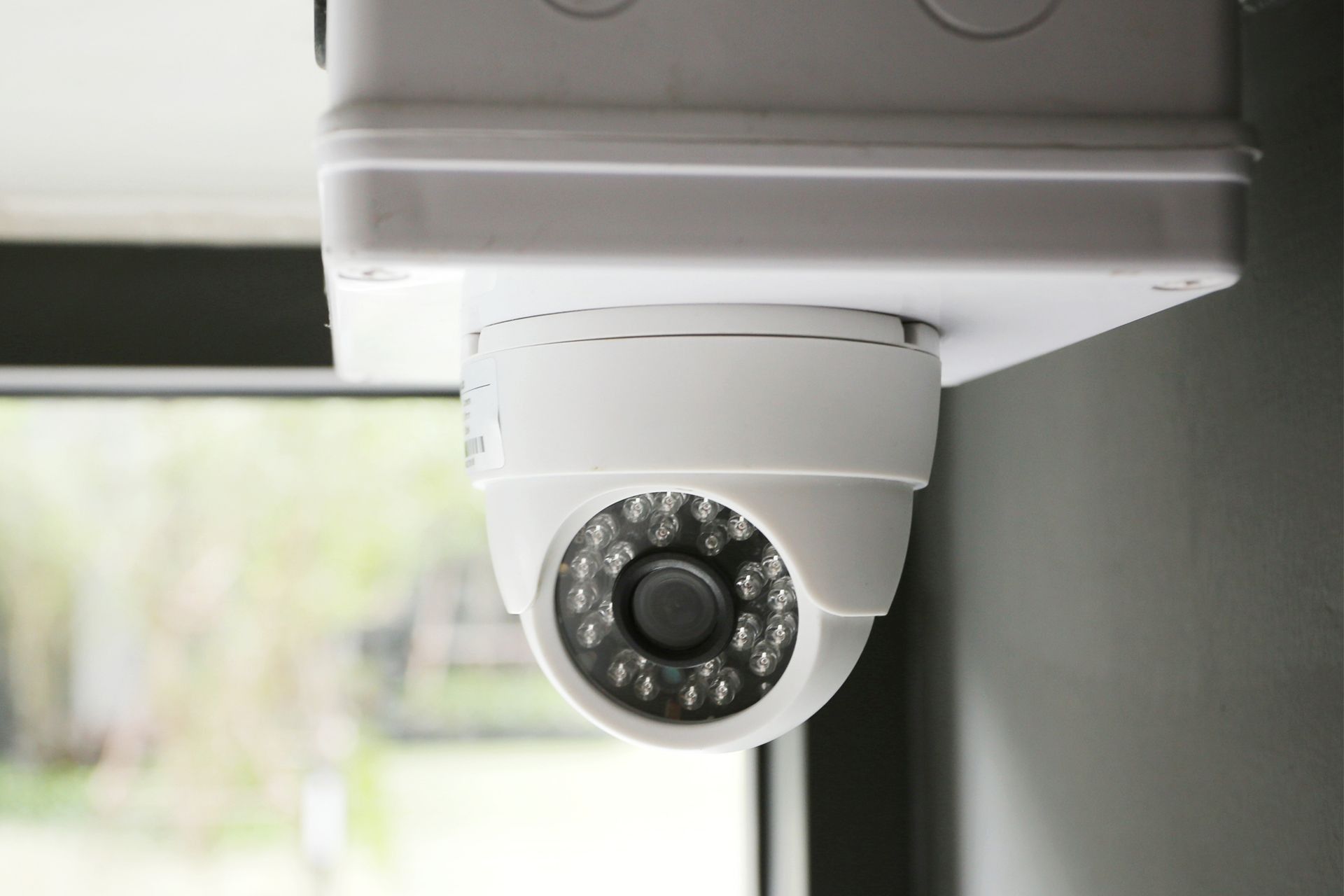 White security camera mounted indoors, facing downward near a window.