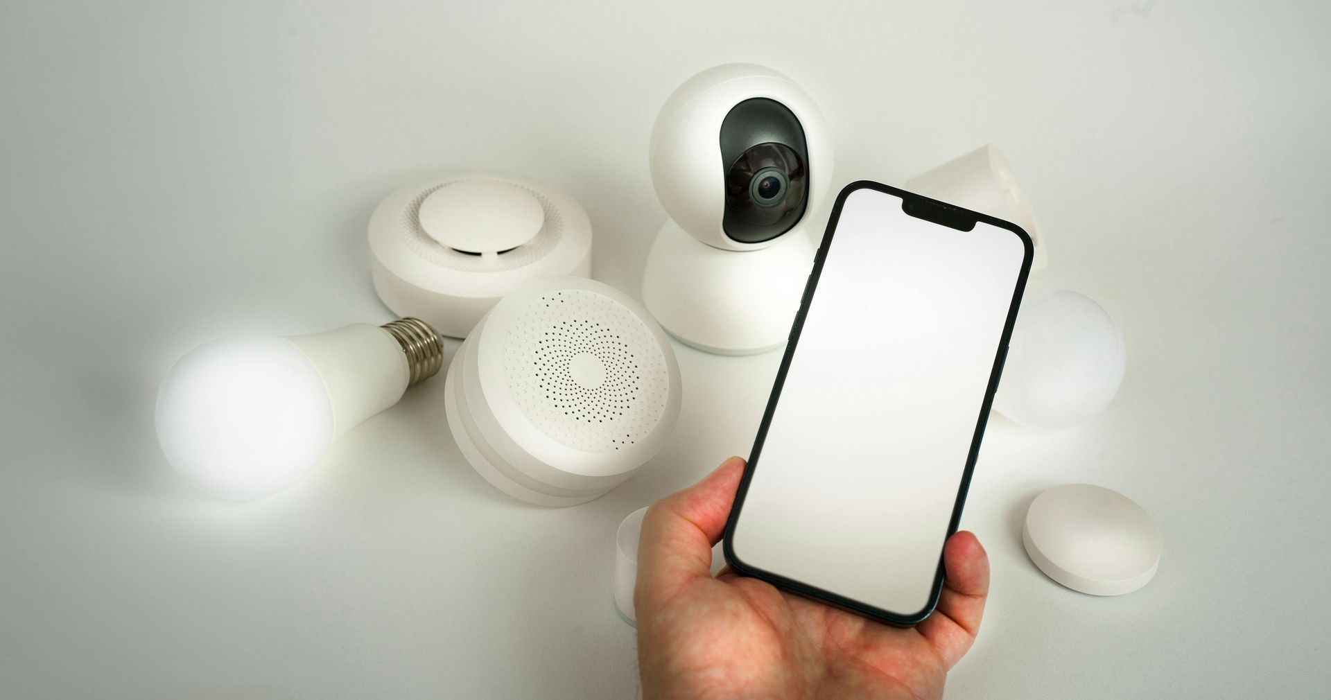 Hand holding a smartphone in front of white smart home devices, including a camera and sensors.