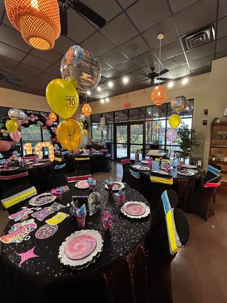 A room with tables and chairs set up for a party with balloons hanging from the ceiling.