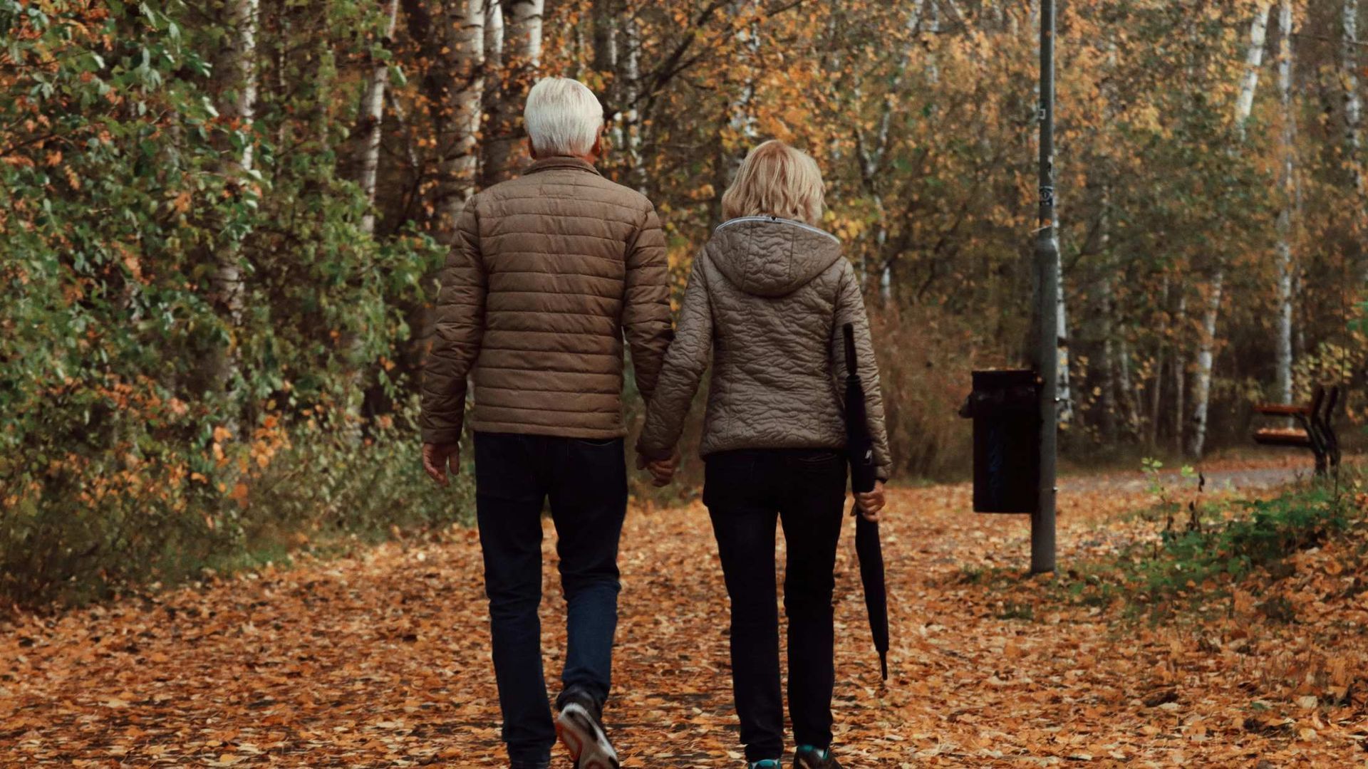 Two people walking away on a leaf-covered forest path in autumn, one carrying an umbrella