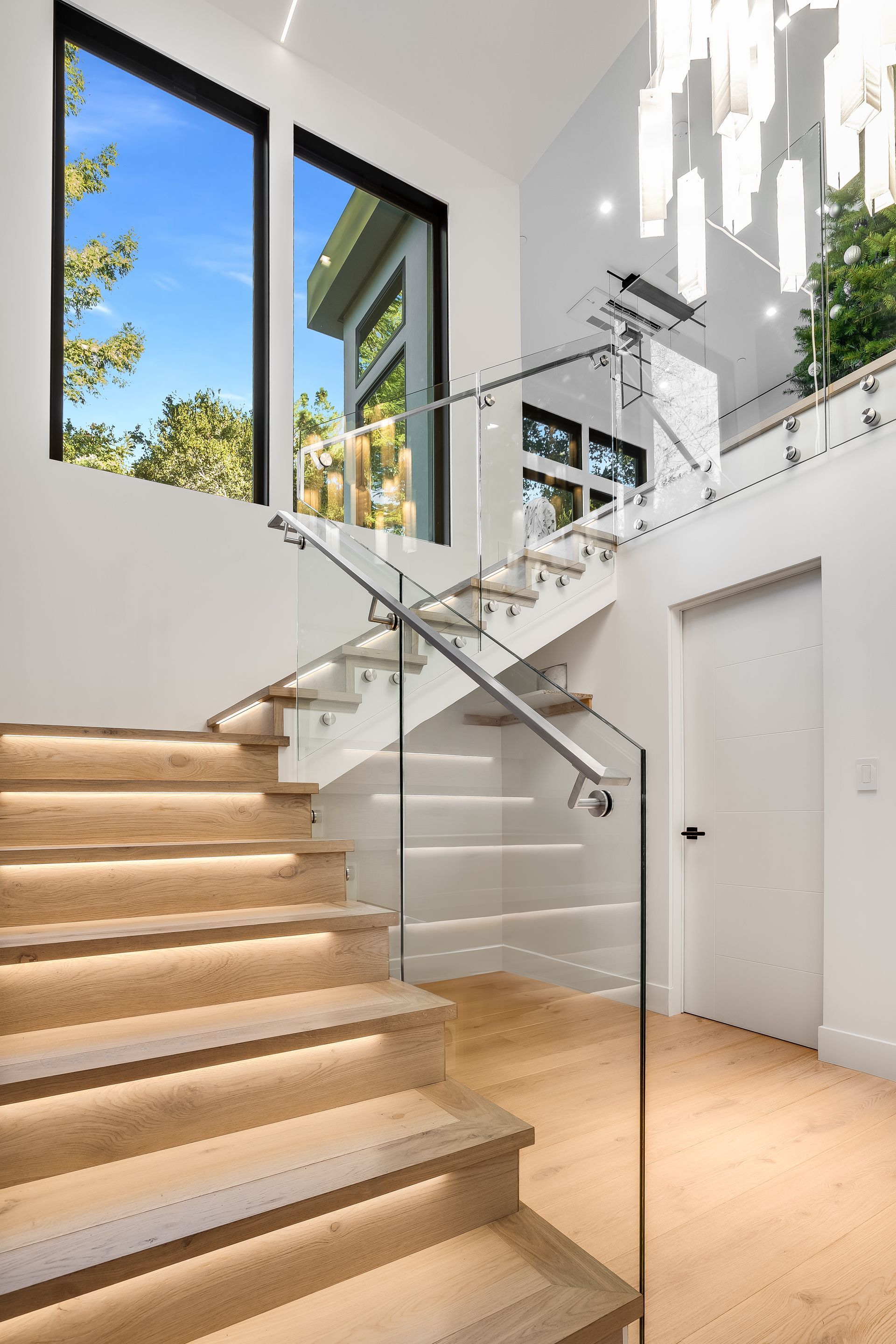 Modern staircase with glass railing, white walls, and large windows in a bright foyer