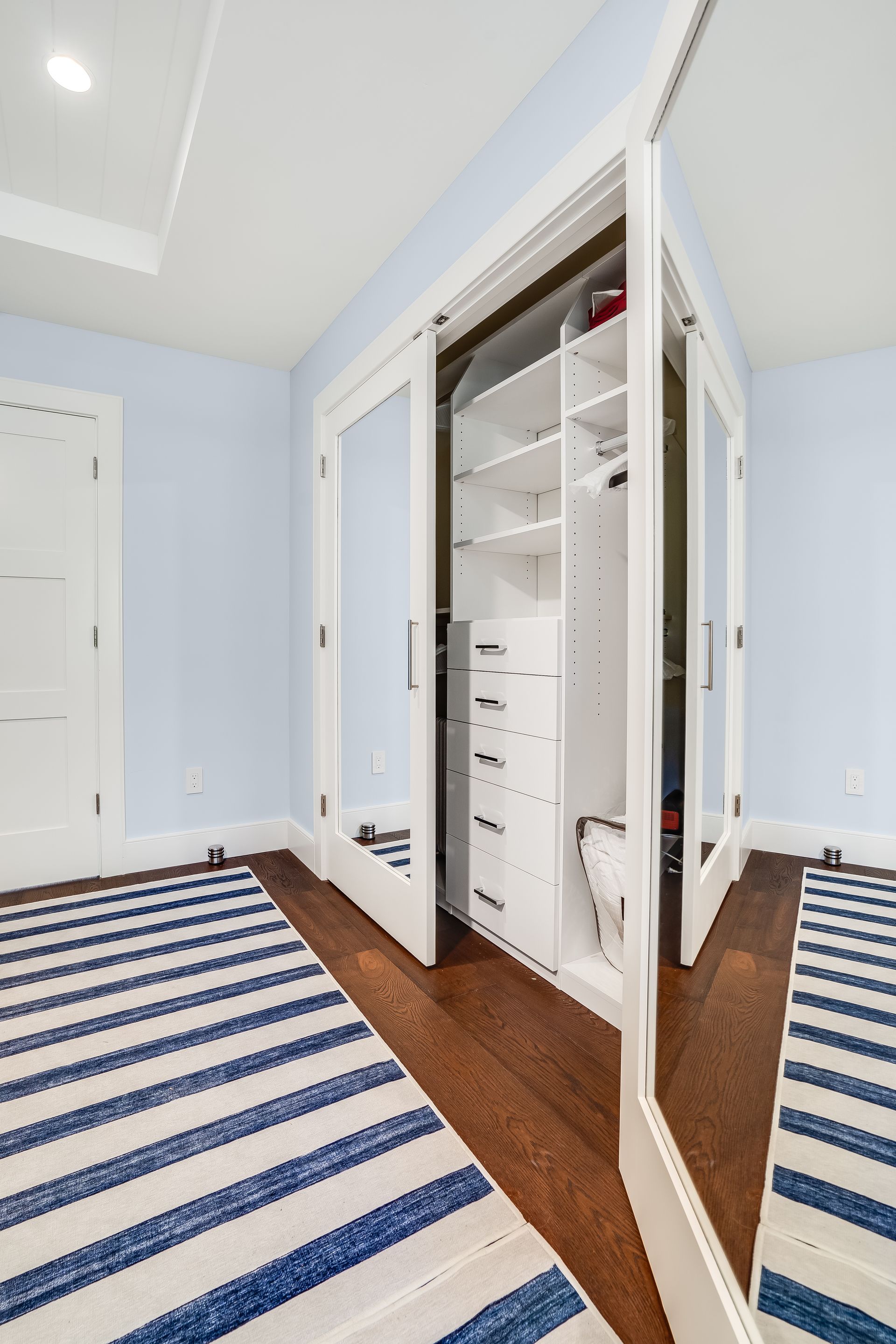 Open closet with white doors and shelves in a light blue bedroom with striped rug and wood floor