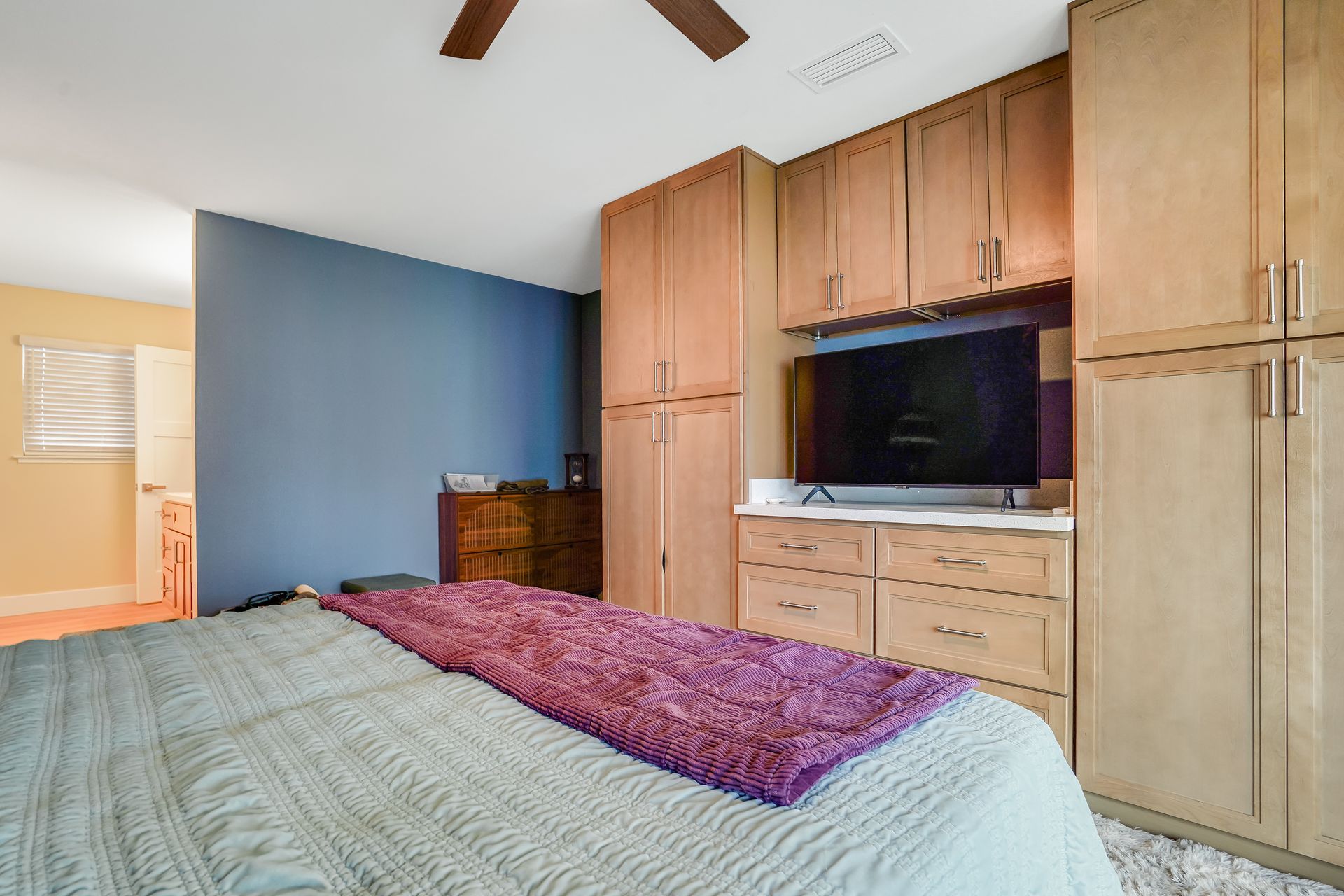 Bedroom with bed, purple blanket, and wood cabinets around a wall-mounted TV