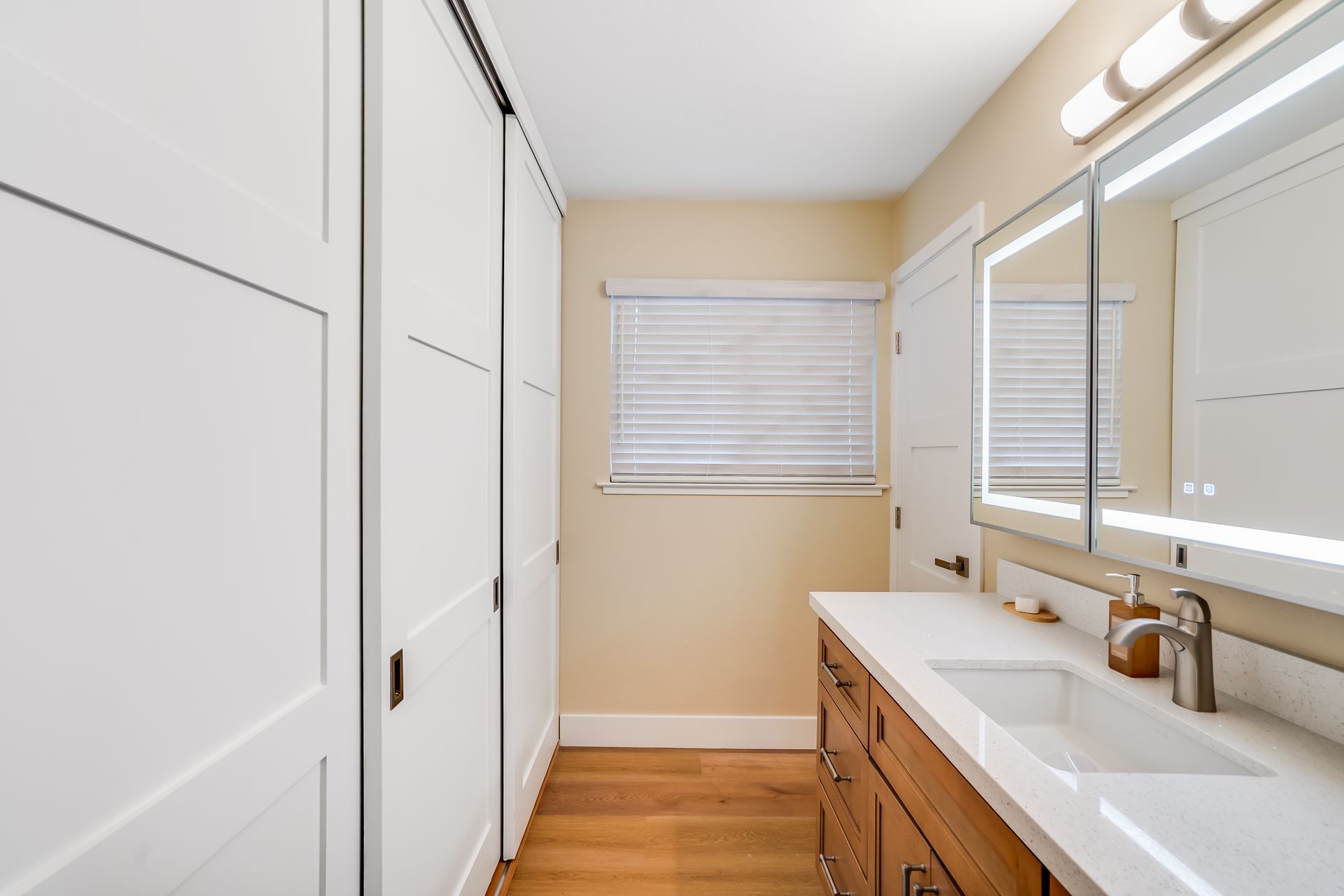 Bright bathroom with white cabinets, wooden vanity, large mirror, and a window with blinds.