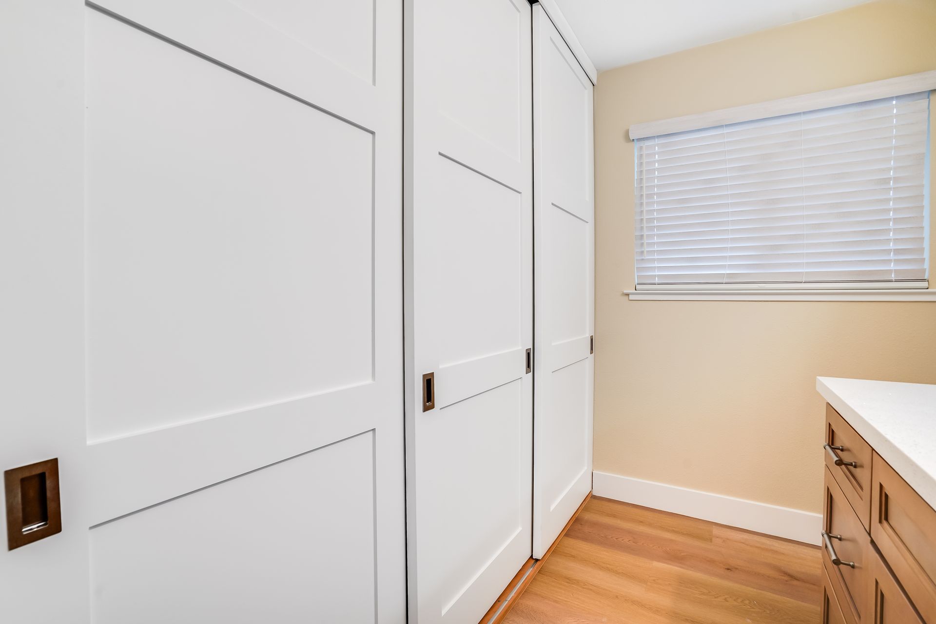 White closet doors in a bright room with wood floor, window blinds, and a wooden cabinet.