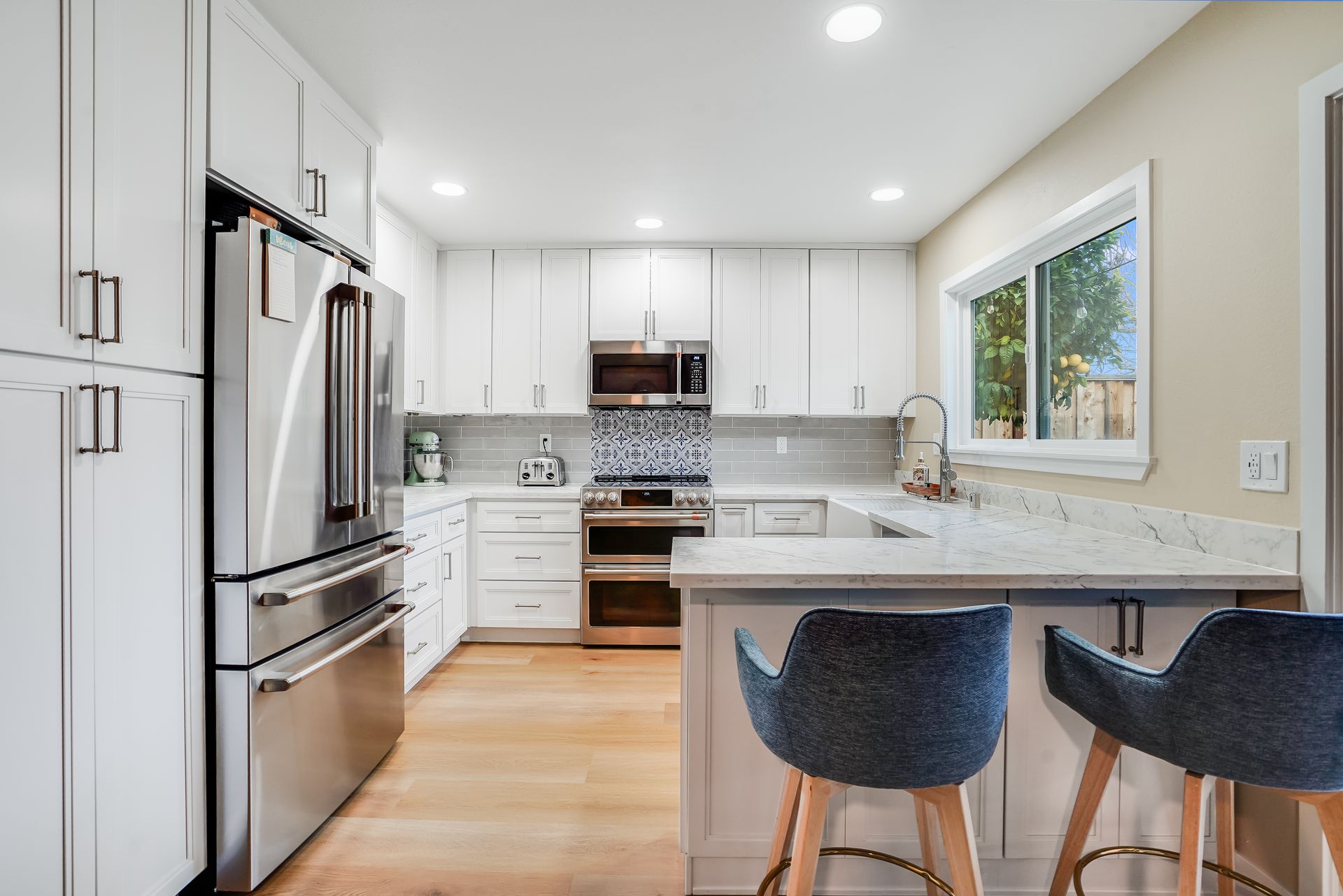 Bright modern kitchen with white cabinets, stainless fridge, and two dark bar stools at a marble island.