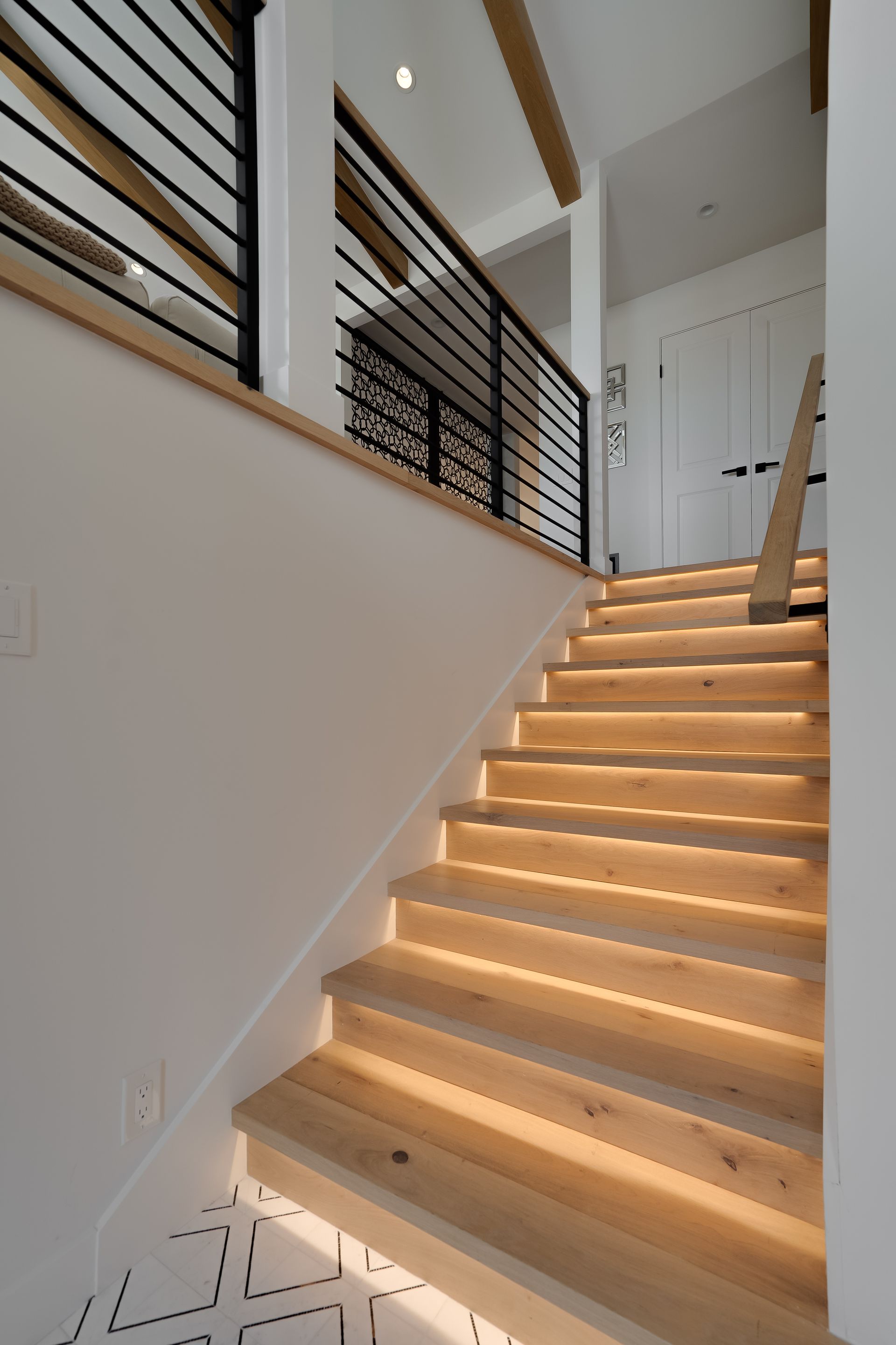 Wooden staircase with under-step lighting, black railing, and white walls.