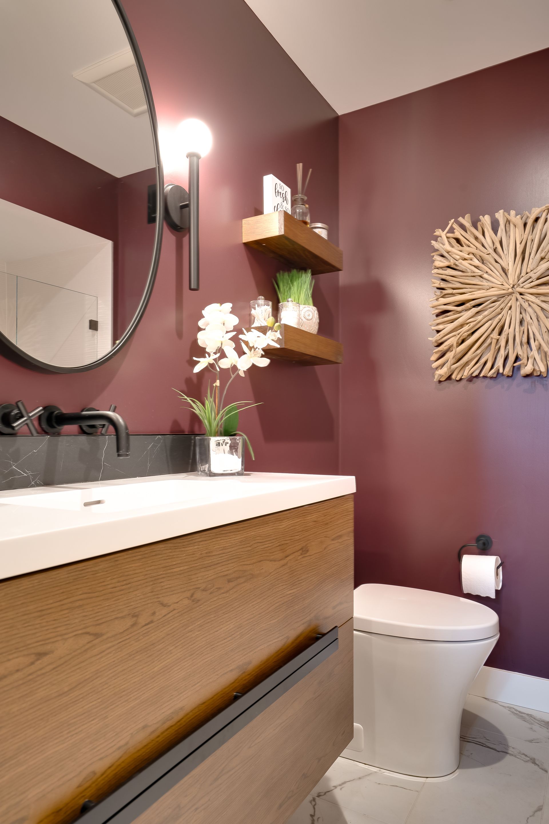 Bathroom with burgundy walls, wooden vanity, floating shelves, and a white toilet.