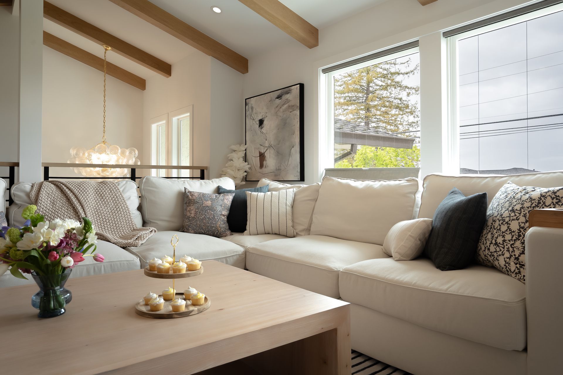 Cozy living room with white sectional sofa, wooden beams, and a view of a bridge. A table holds treats and flowers.