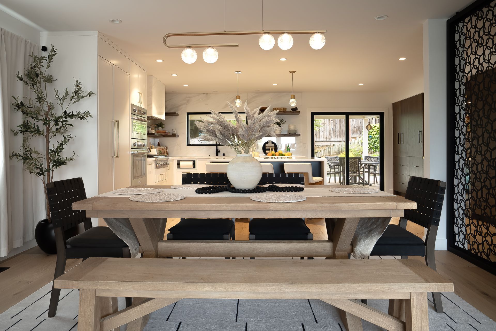 Dining room with wooden table, black chairs, and modern light fixtures; kitchen in the background.