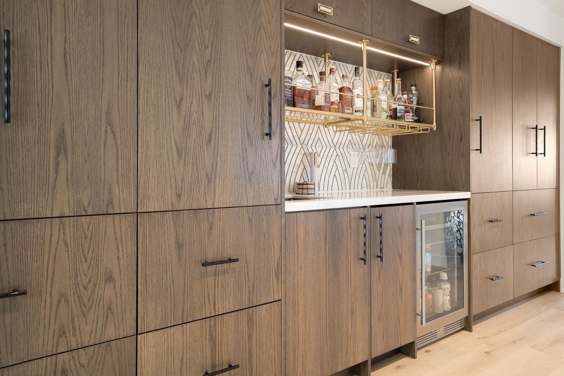 Modern home bar with wood cabinets, a small refrigerator, and shelves with liquor bottles.