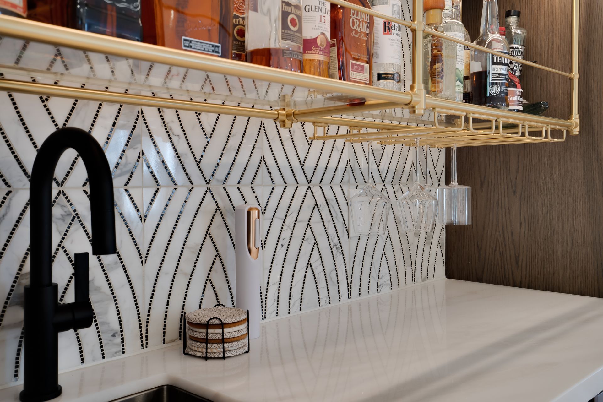 Black faucet, white countertop, diamond-patterned tile backsplash, gold shelving with liquor bottles, and bar accessories.