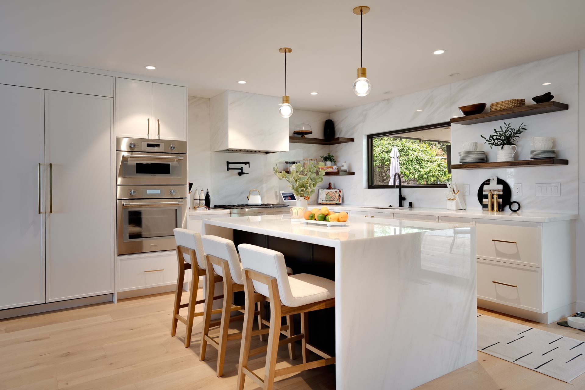 Modern white kitchen with island and bar stools, featuring stainless steel appliances, and a window.
