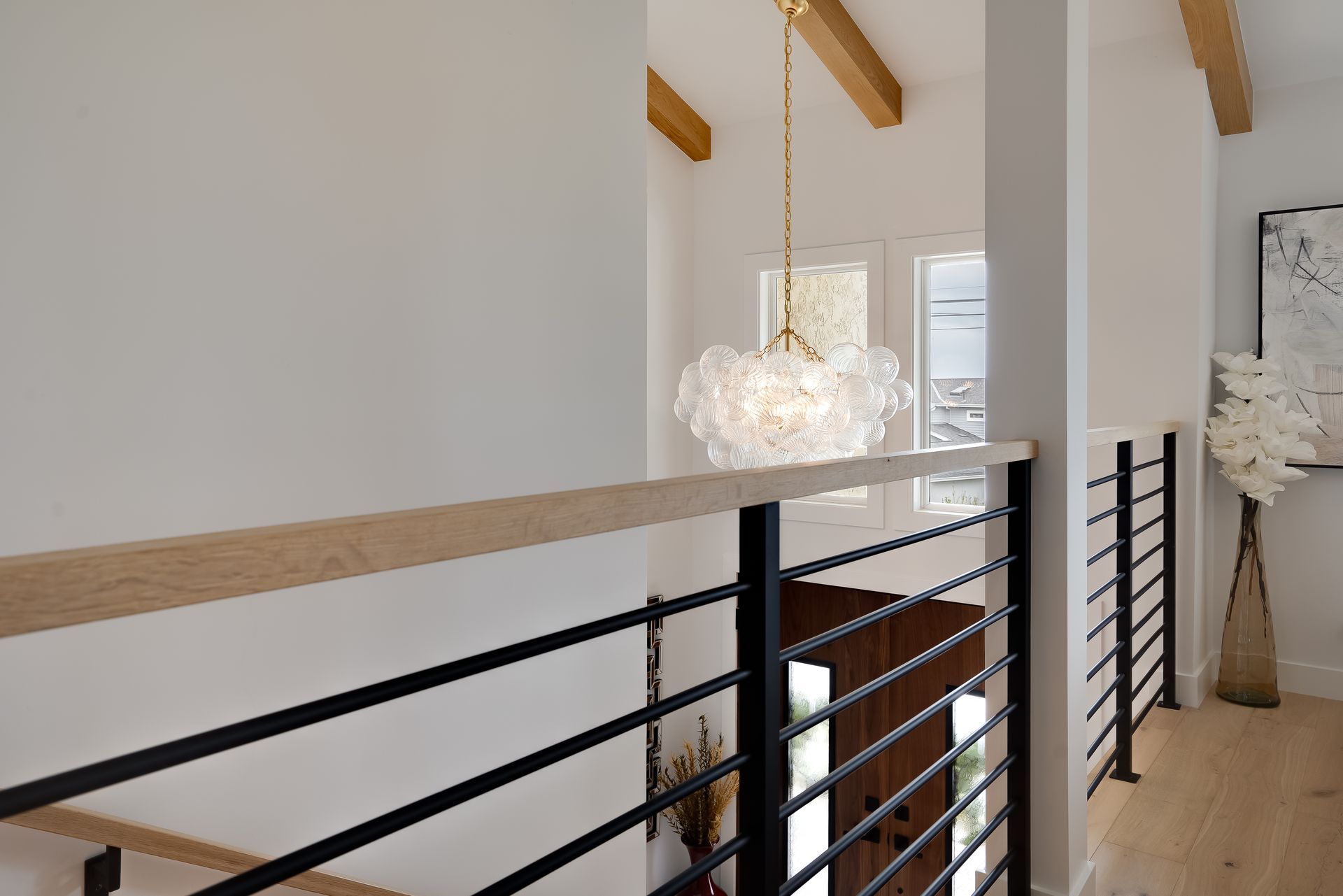 Hallway with gold chandelier, wooden beams, black metal railing, white walls, and wooden floor.