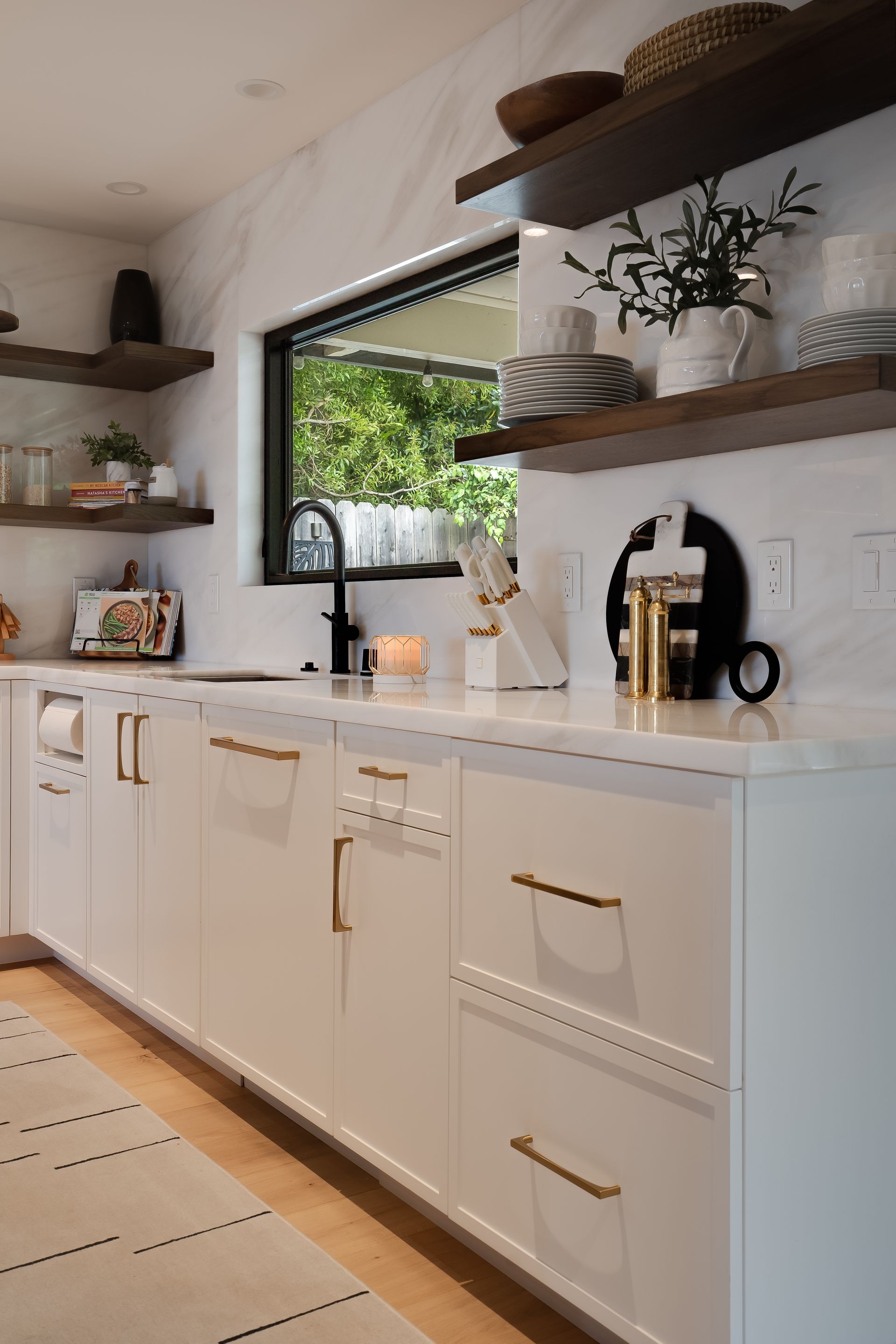 Modern white kitchen with gold hardware, open shelving, and a window overlooking greenery.