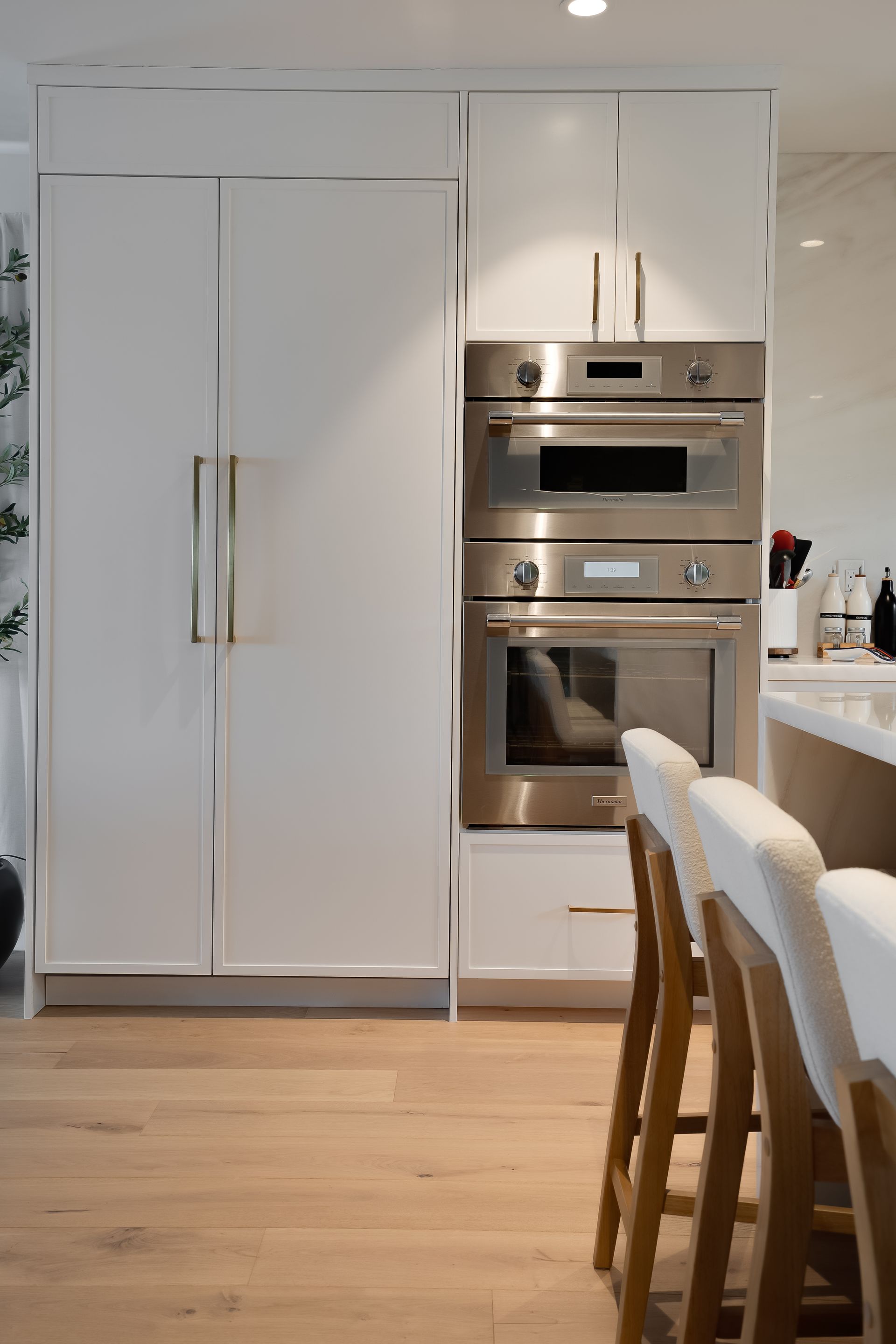 White kitchen with refrigerator and stainless steel ovens. Bar stools in foreground.