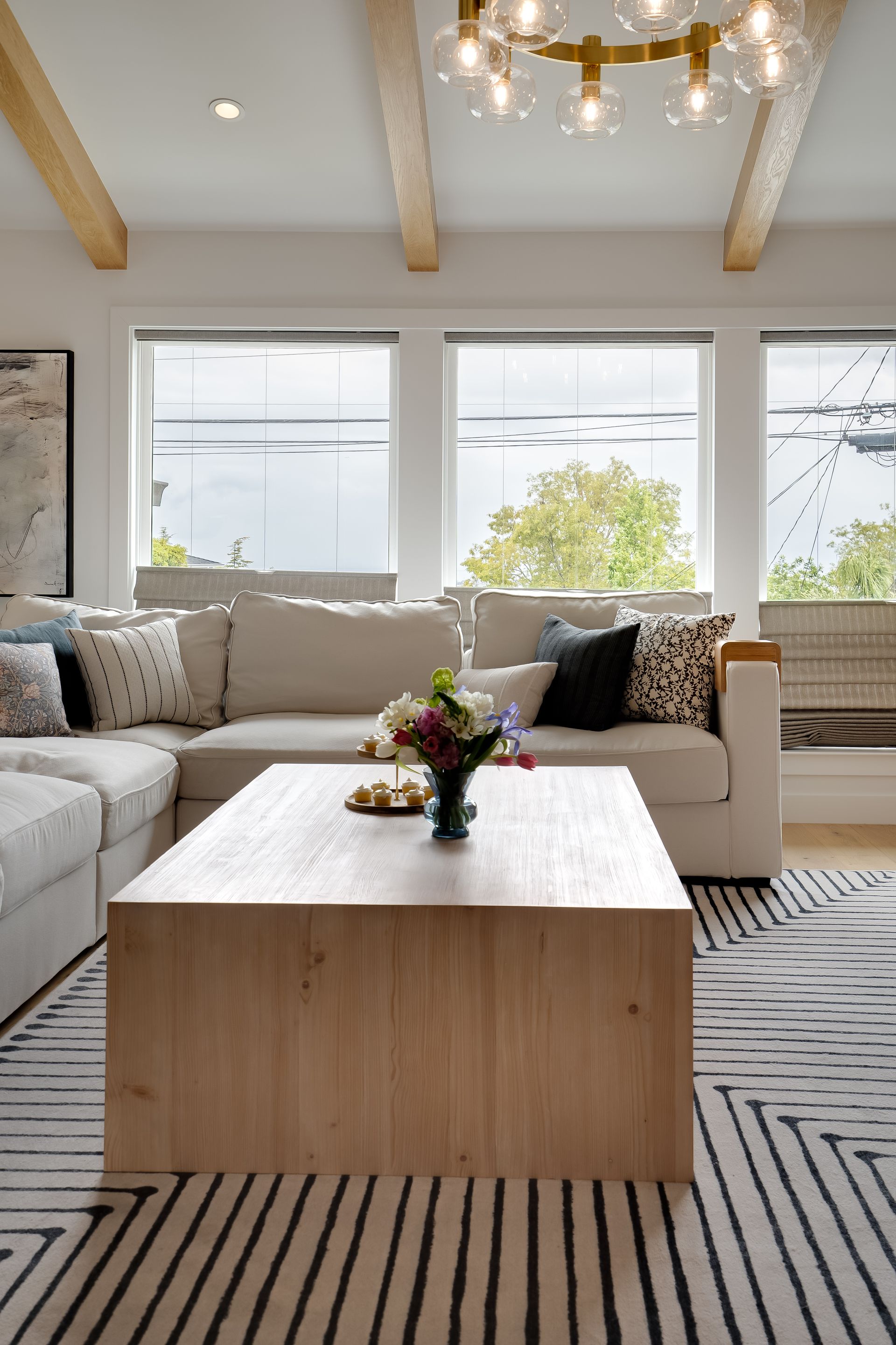 Living room with beige sectional, wooden coffee table, and large windows. Black and white patterned rug.