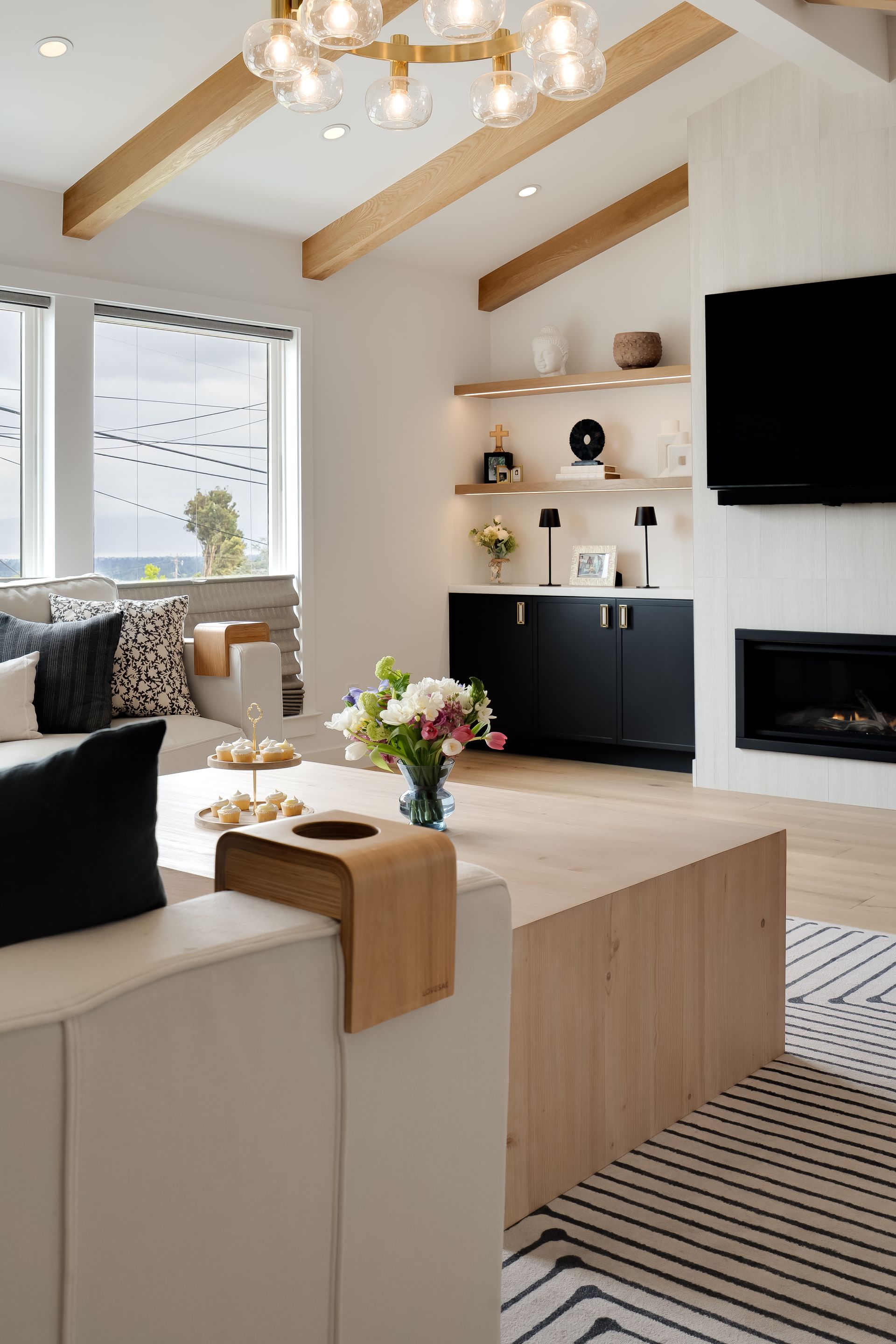 Modern living room: white walls, wooden beams, black cabinets, fireplace, TV, beige couch, and neutral rug.