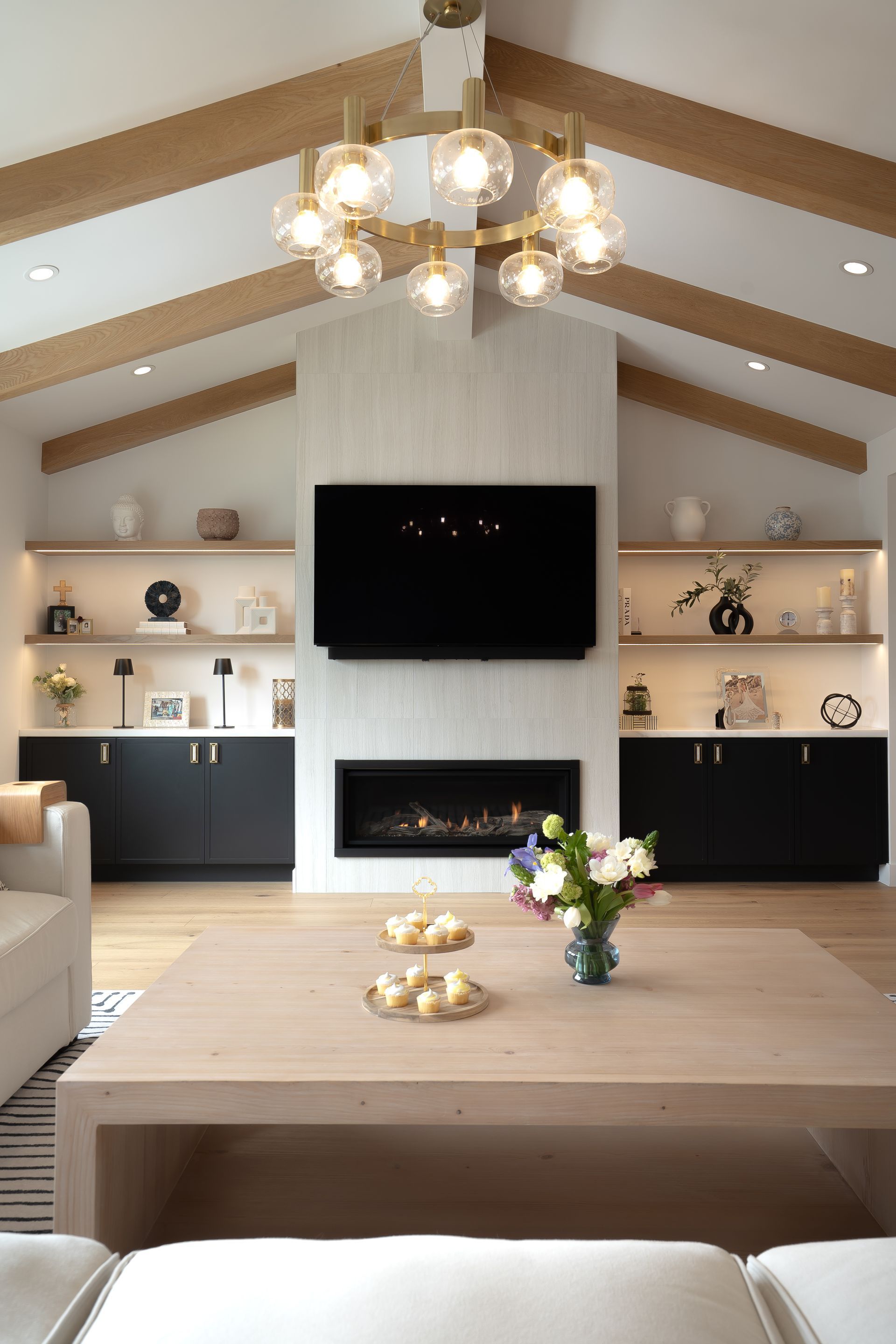 Living room with a fireplace, TV, and decorative shelving. Features a light wood coffee table and a modern chandelier.
