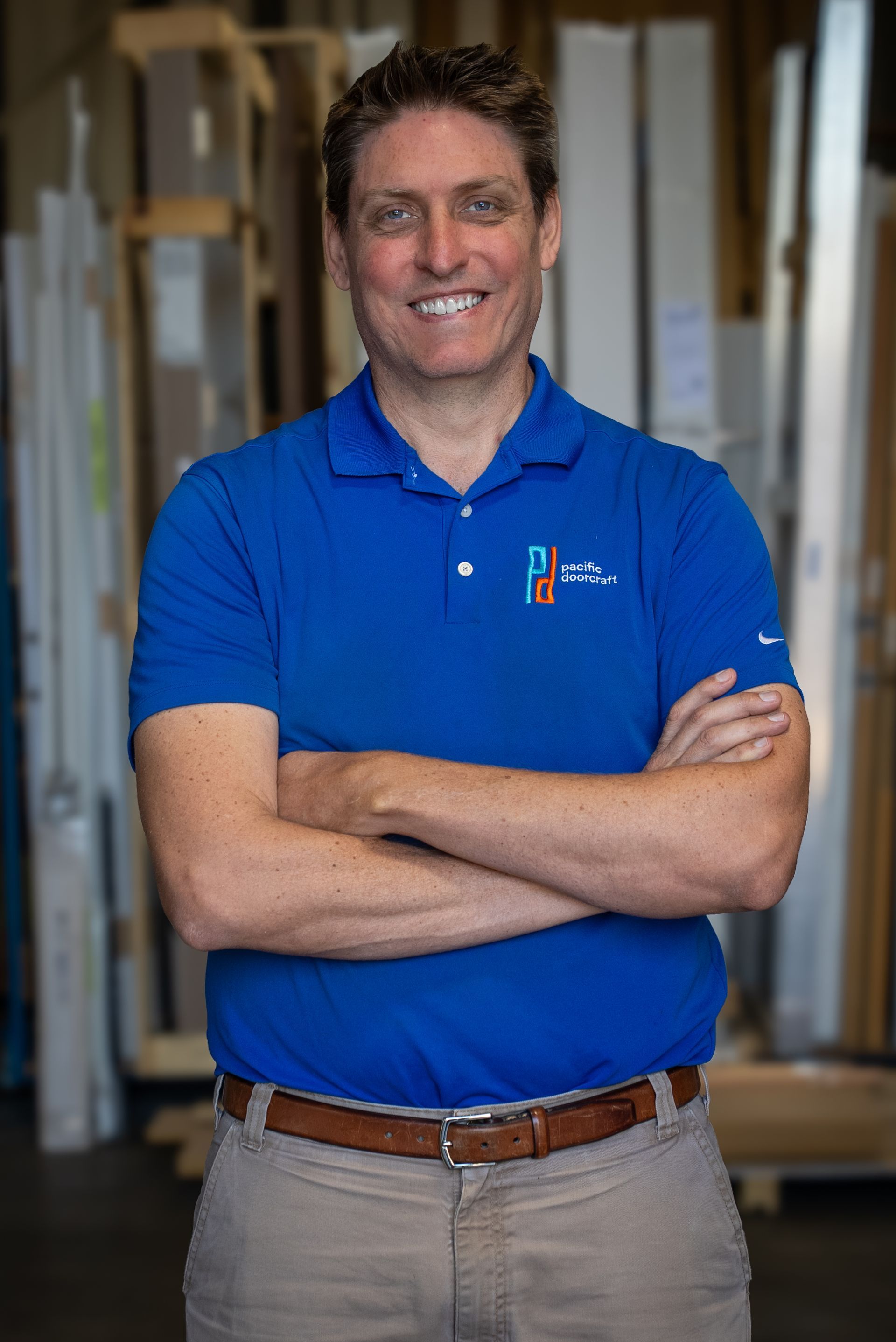 Smiling man in a blue polo shirt standing with arms crossed in a workshop or store aisle.