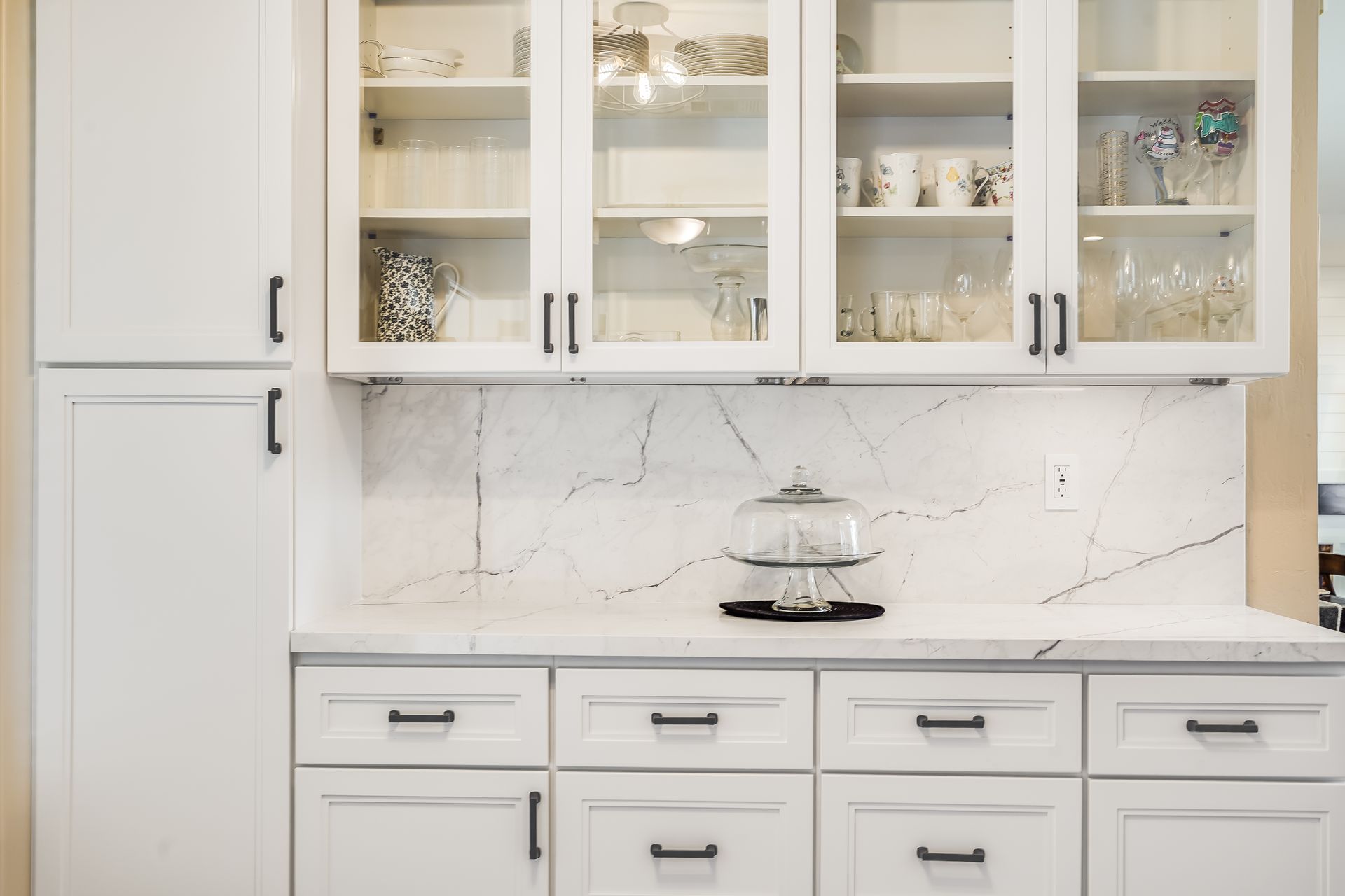 White kitchen with glass-front cabinets, marble backsplash, and a cake stand on the countertop