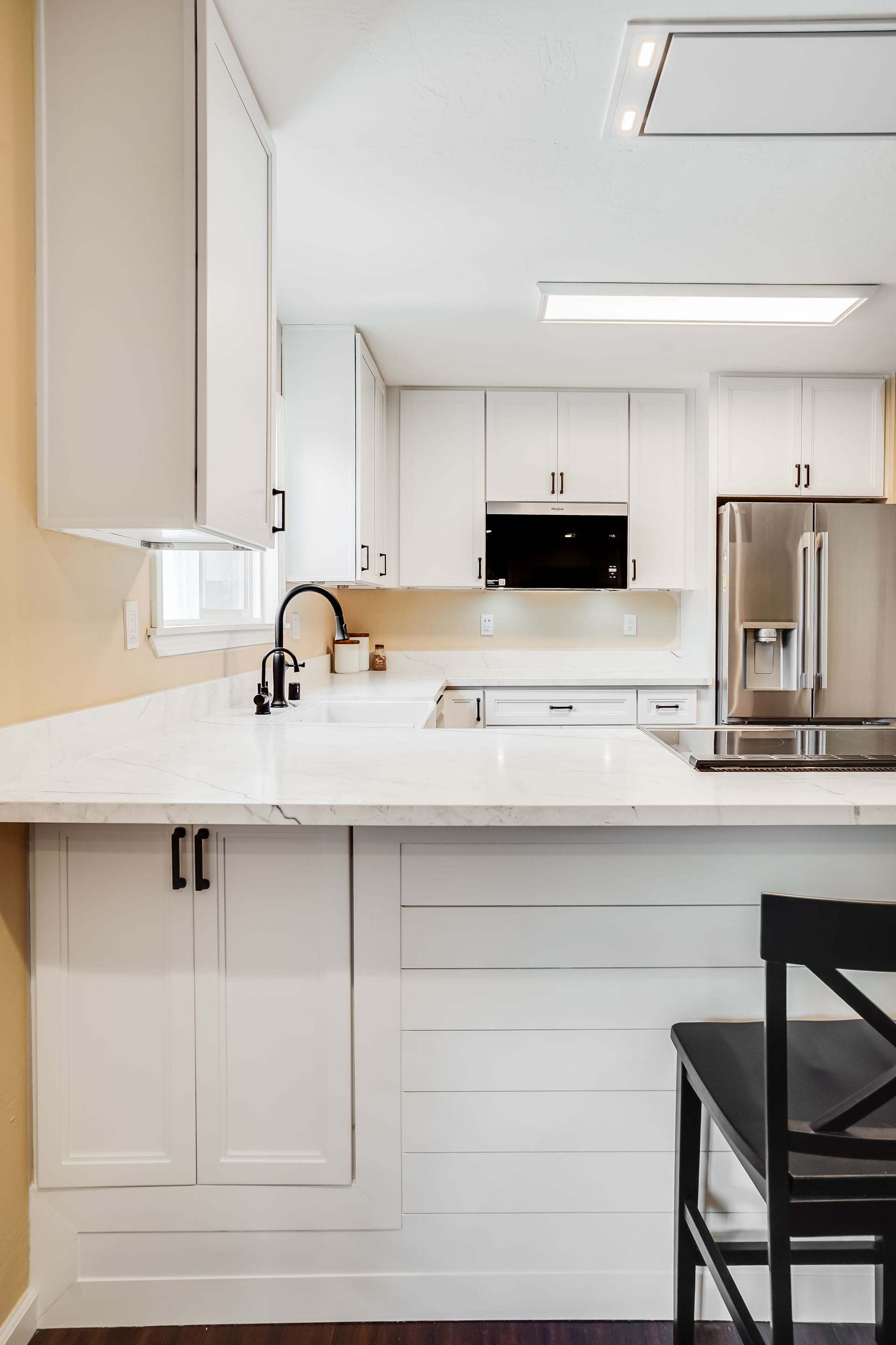 Bright white kitchen with island, stainless steel refrigerator, and black stool under pendant lights