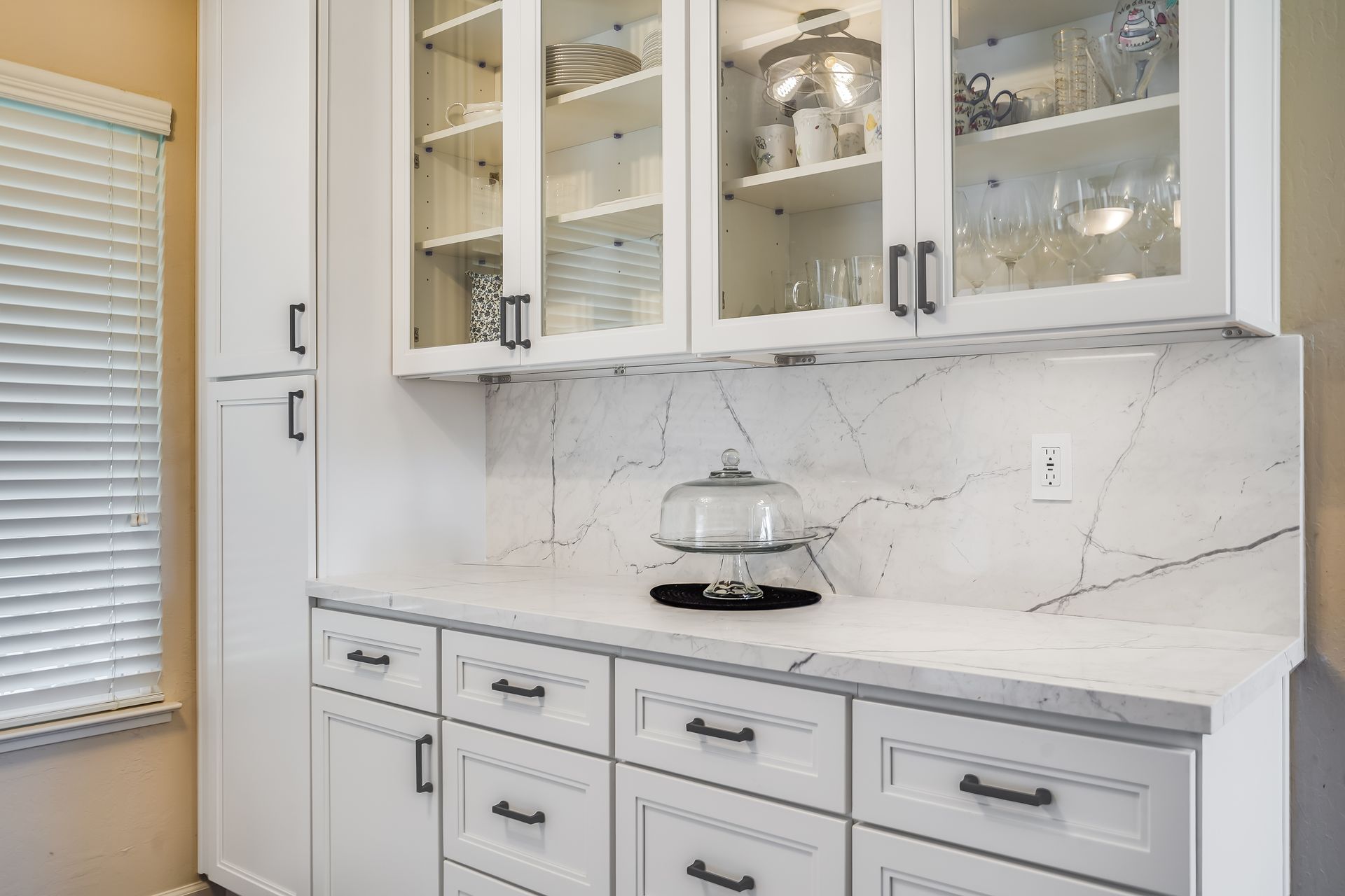White kitchen with glass-front cabinets, marble backsplash, and a countertop mixer.
