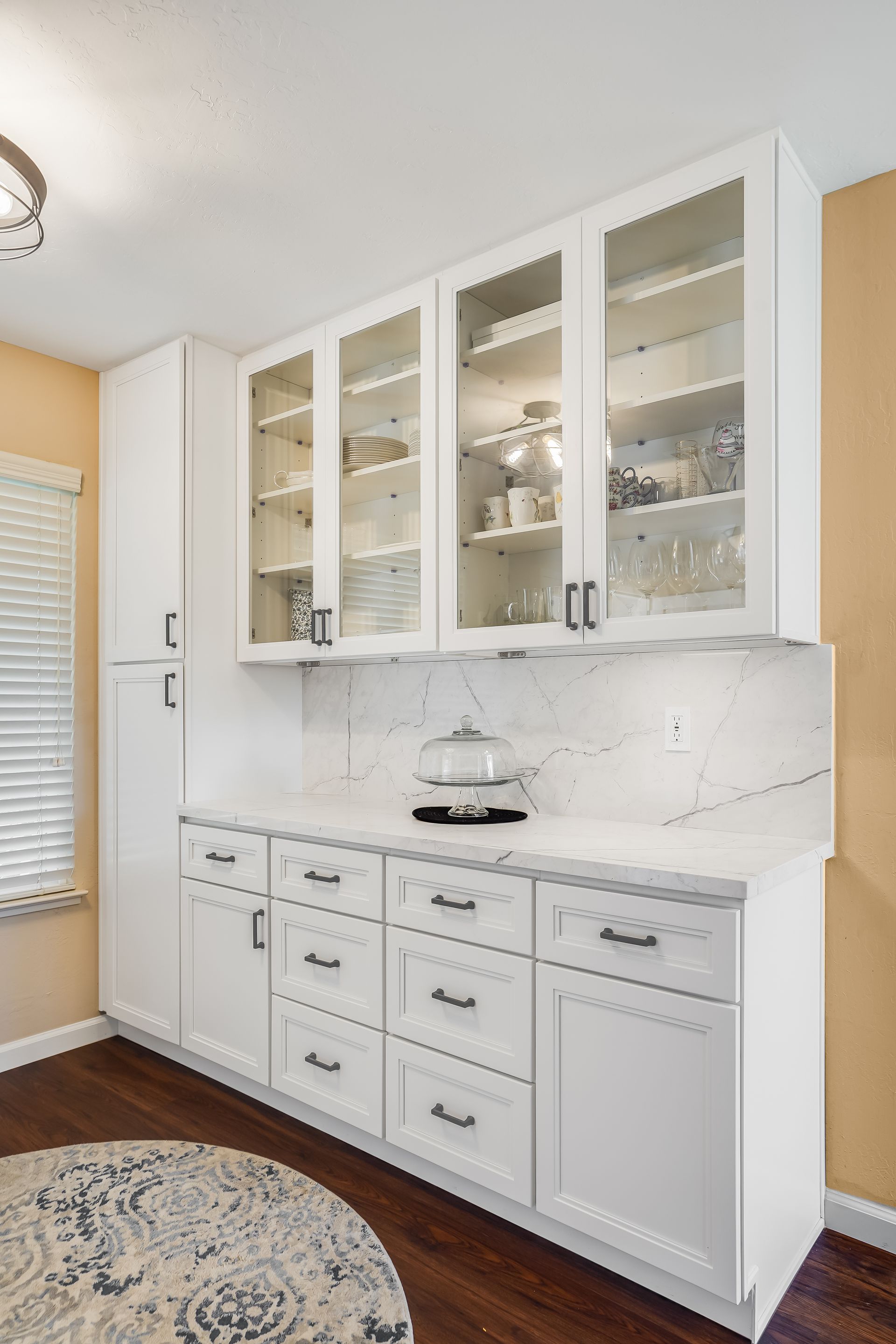 White built-in kitchen hutch with glass cabinets, drawers, and marble countertop against tan walls