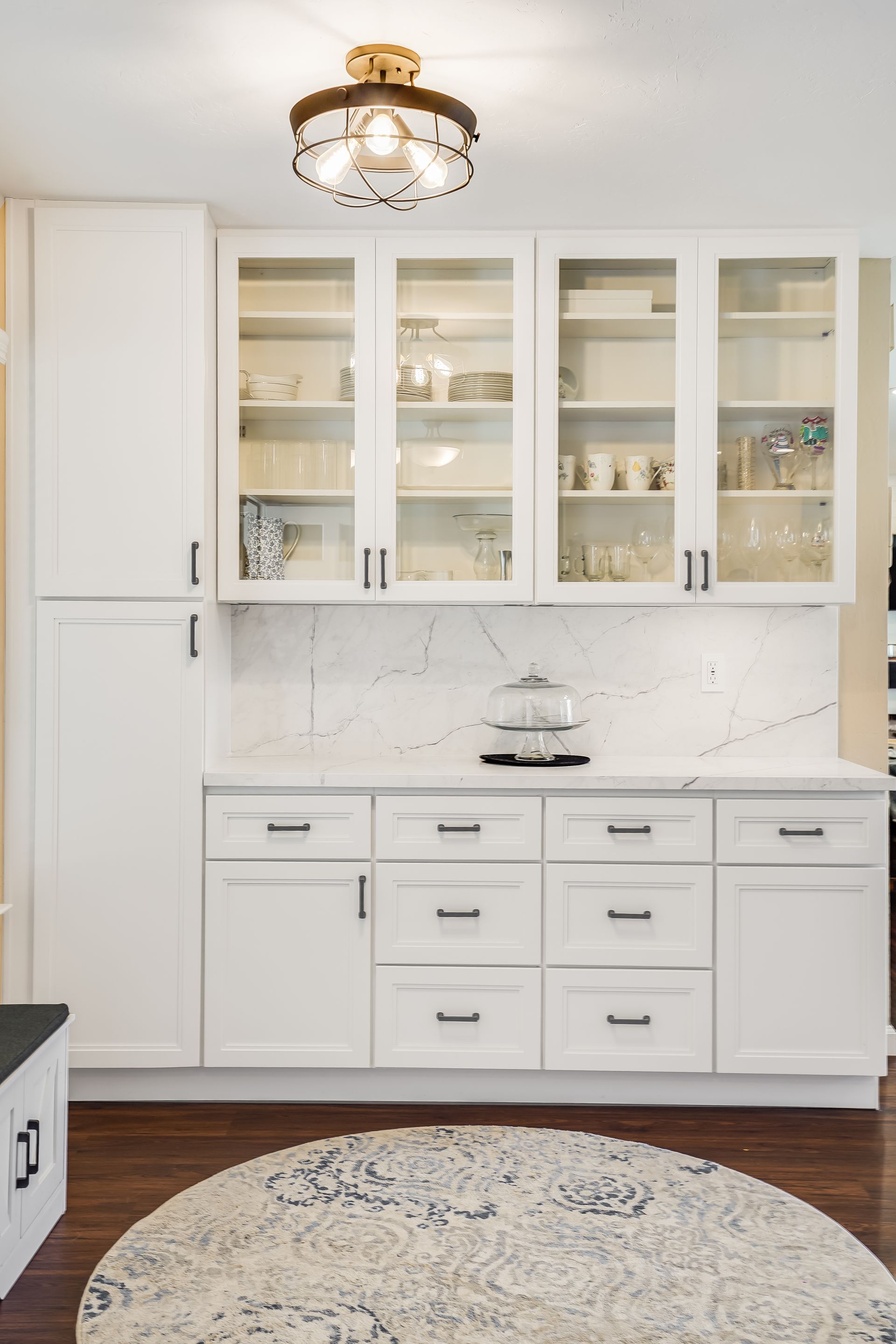 White built-in kitchen cabinets with glass doors, marble backsplash, and round rug on dark wood floor