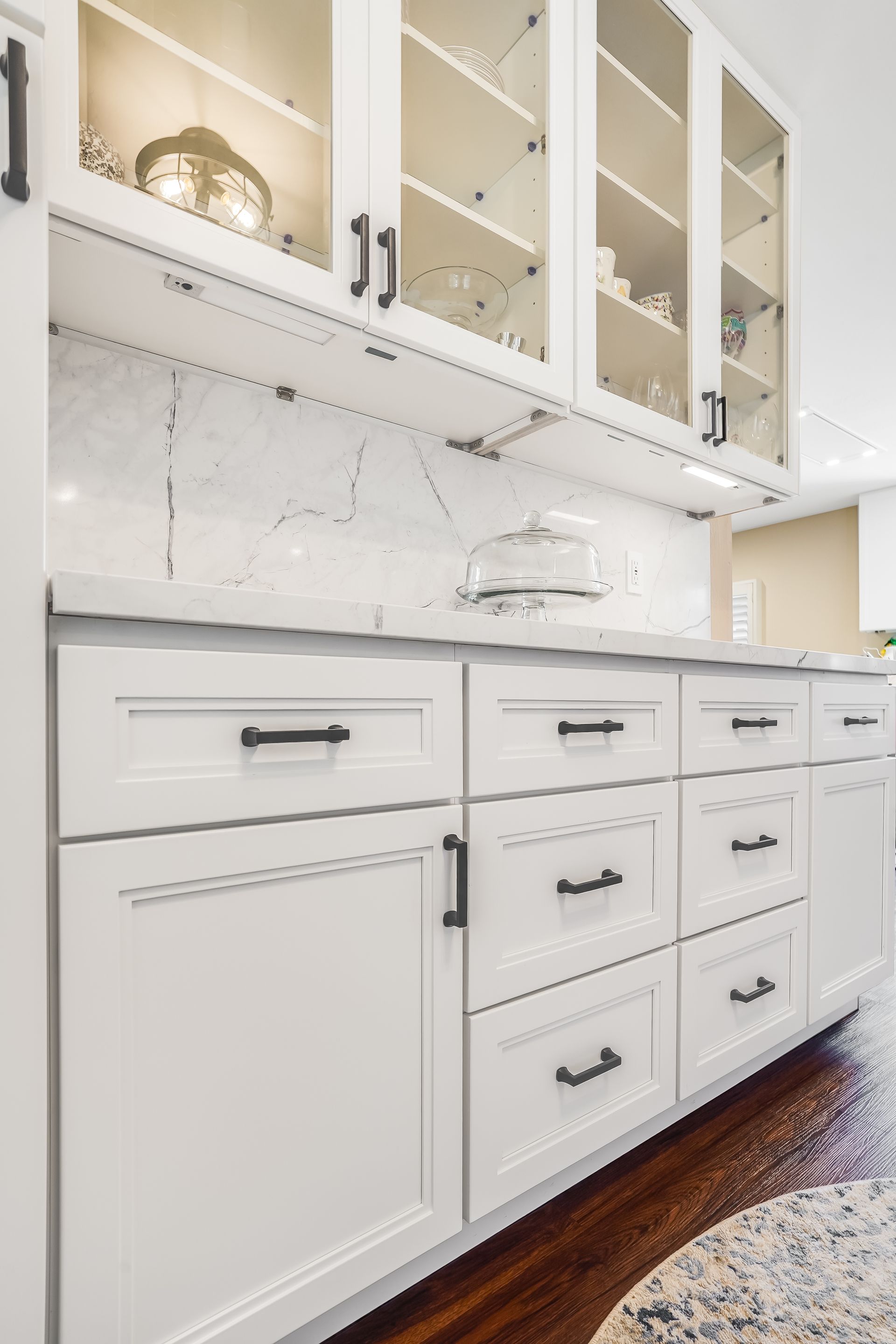 White bathroom vanity with cabinets, drawers, and glass-front upper cabinets against a marble wall.