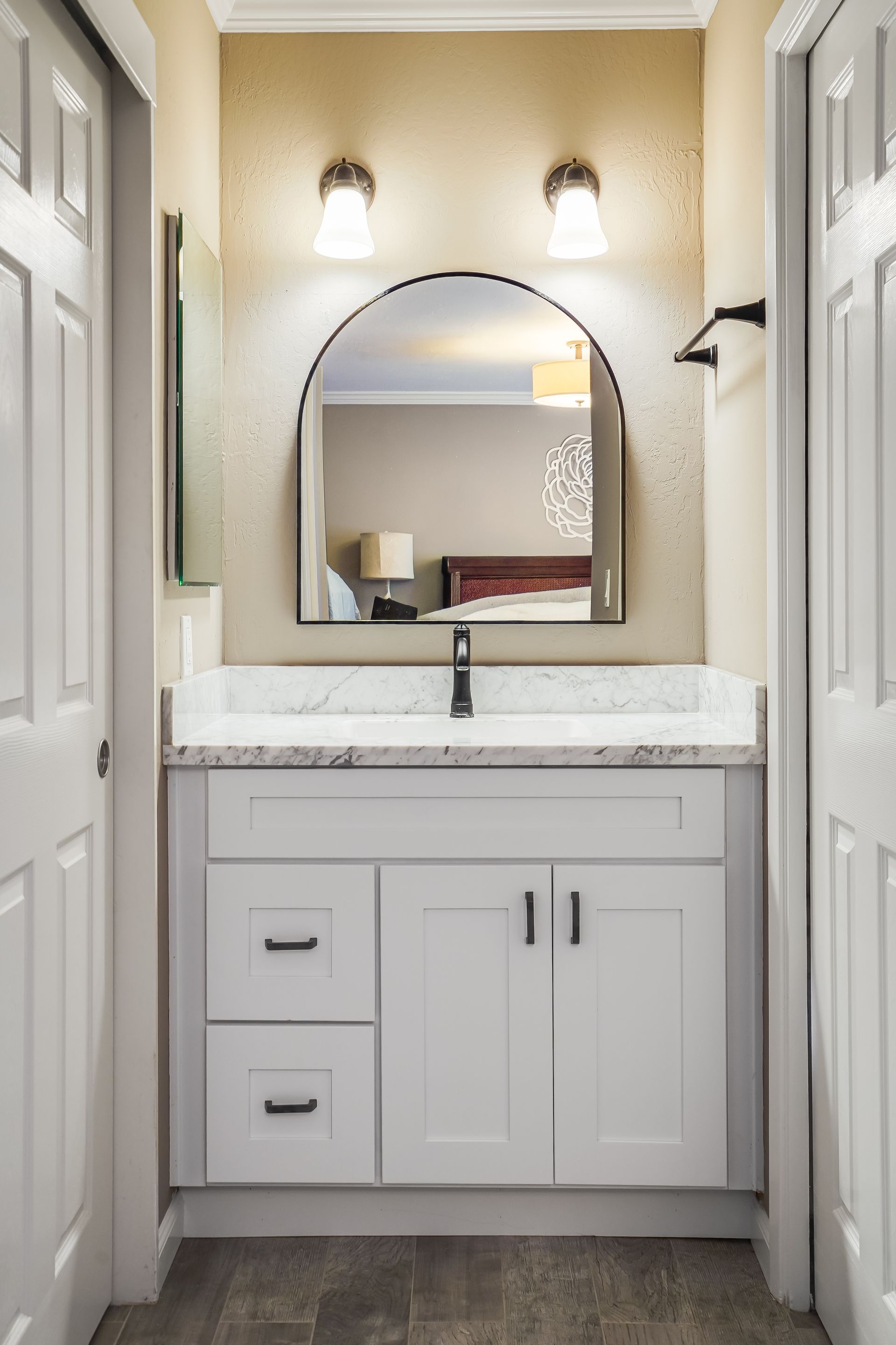 Small white bathroom vanity with marble countertop, arched mirror, and two wall sconces between closet doors
