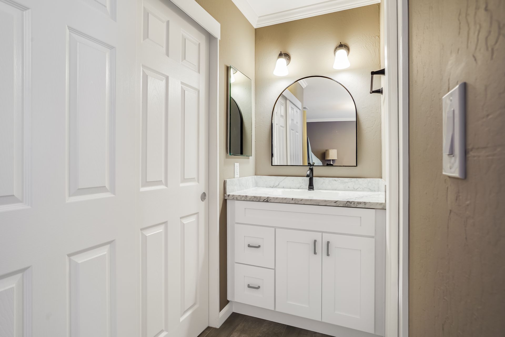 Bright bathroom vanity with white cabinets, arched mirror, and wall sconces beside a closed door.