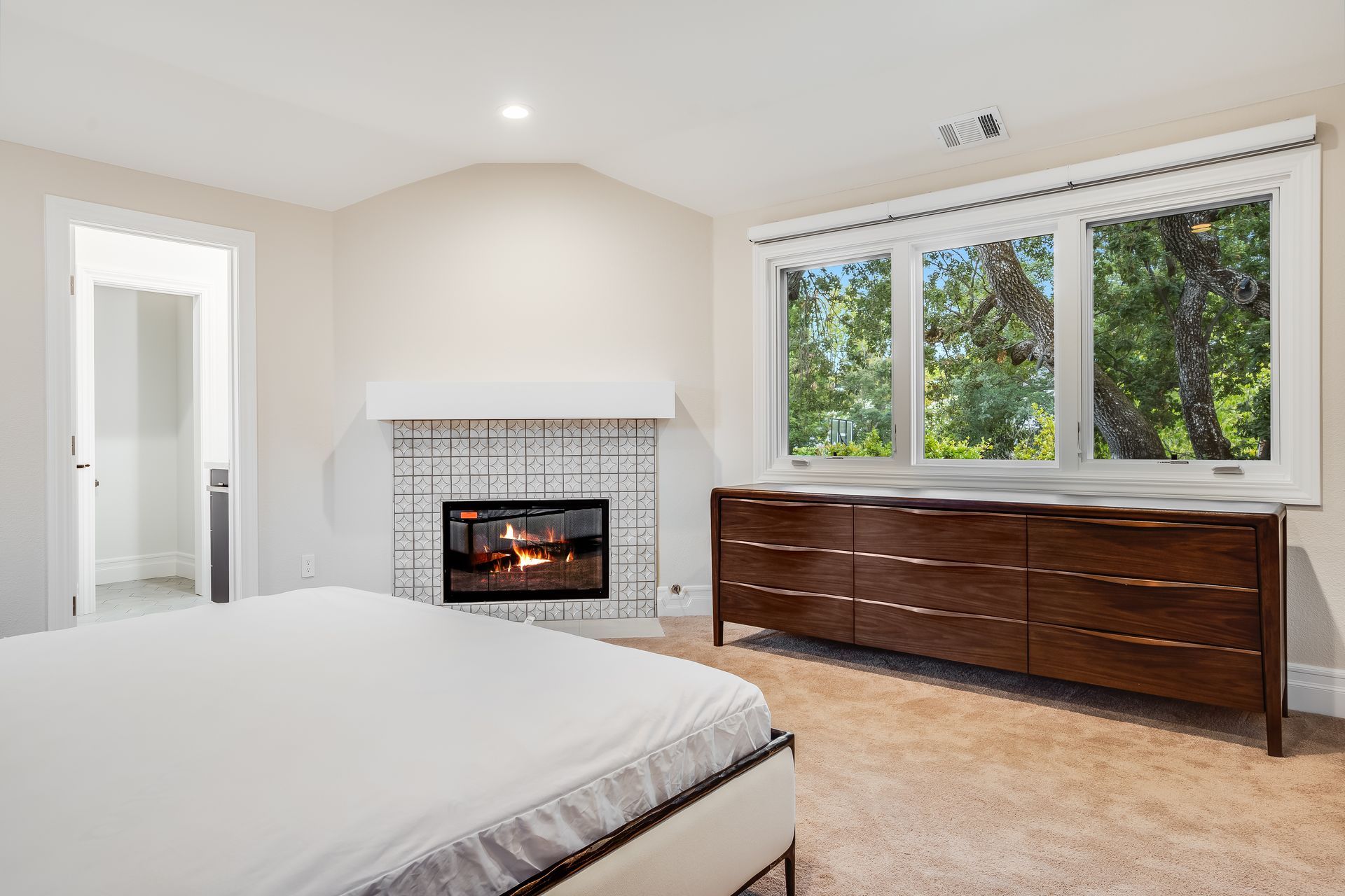 Bright bedroom with bed, tiled fireplace, long dresser, and large windows overlooking trees