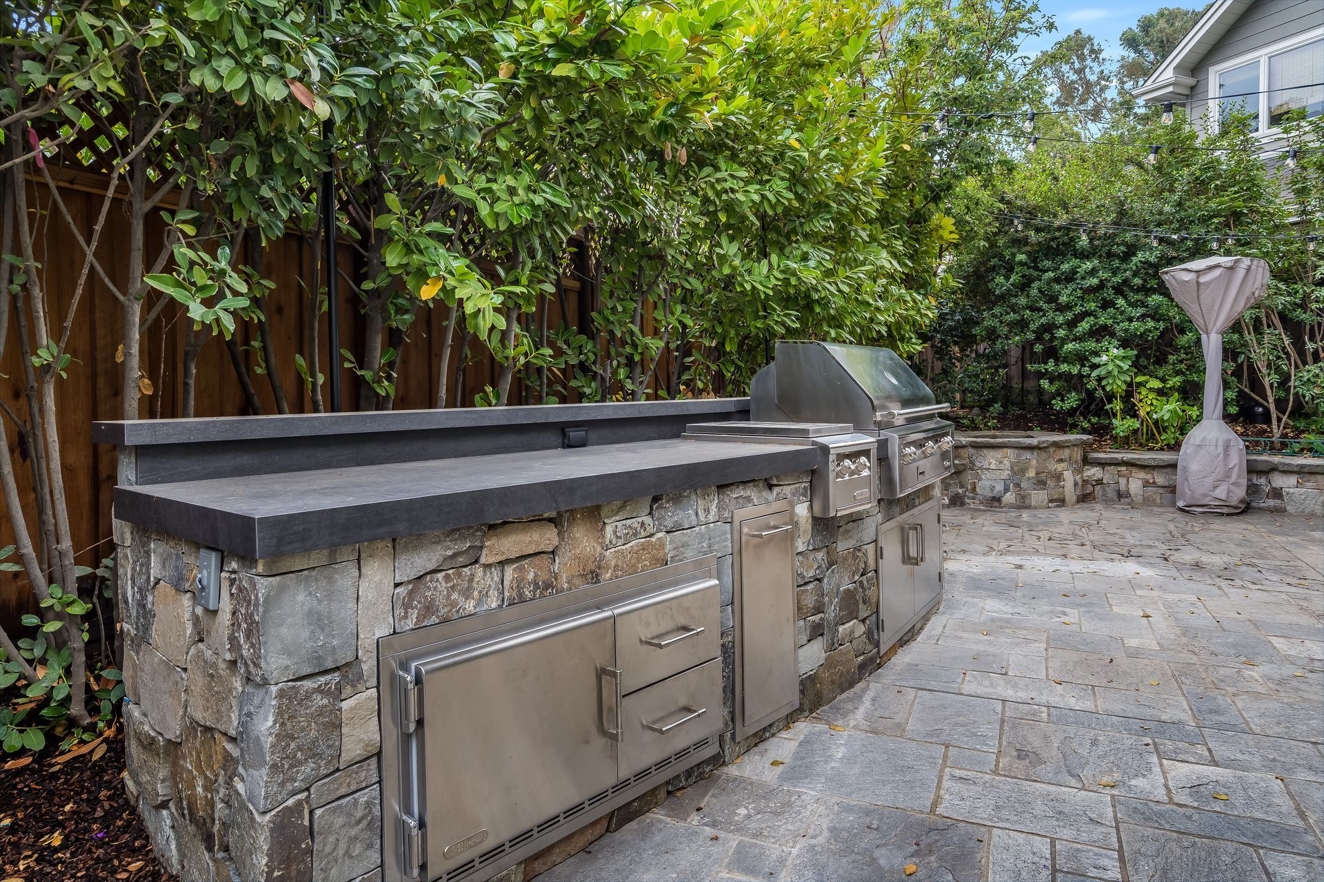 Outdoor stone patio with built-in grill and cabinets beside a wooden fence and greenery