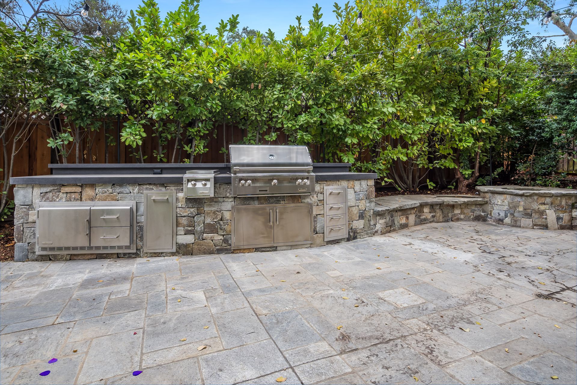 Outdoor stone patio with built-in stainless steel grills and a lush green hedge backdrop
