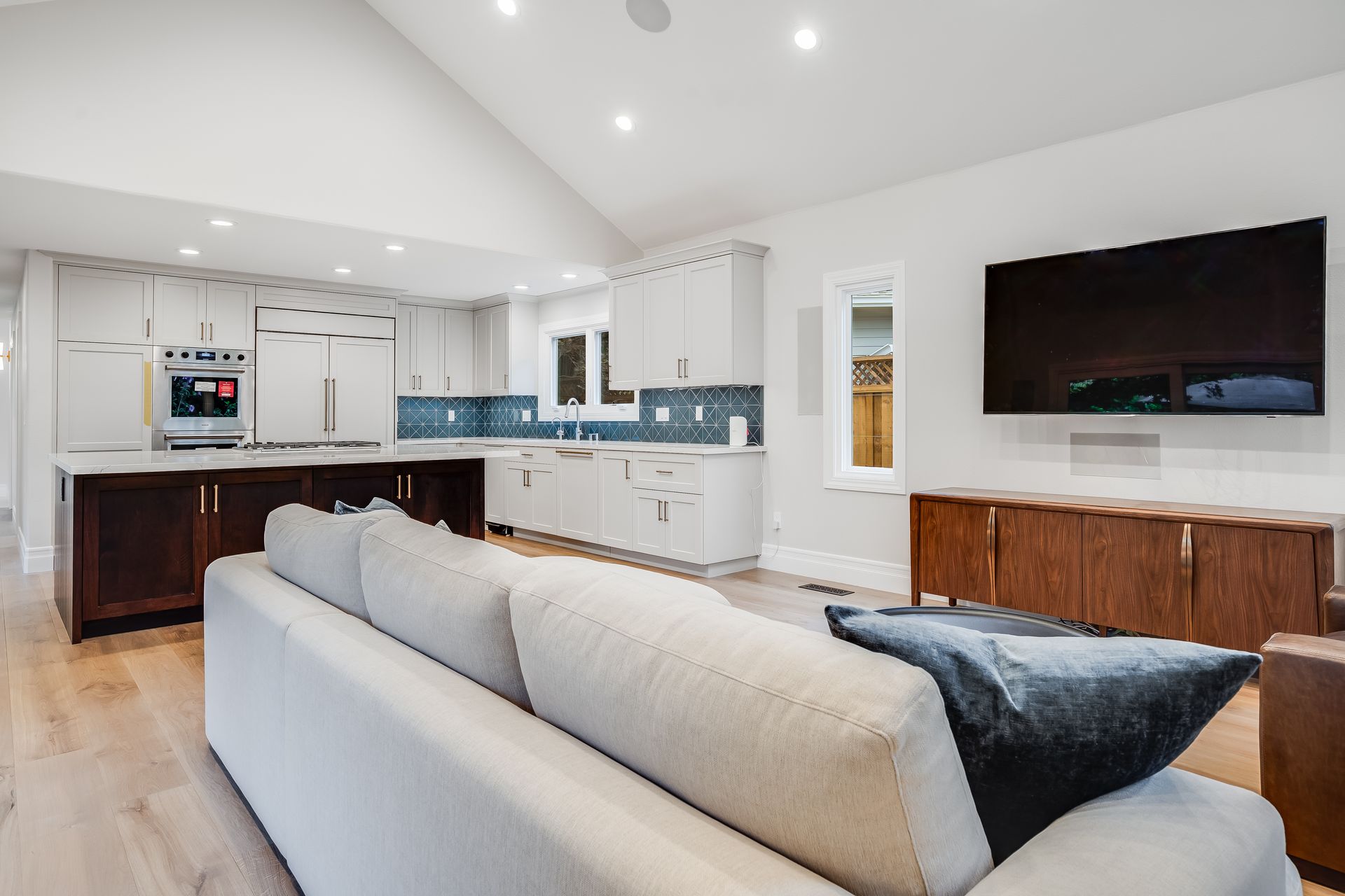 Bright modern living room with gray sofa, wall-mounted TV, and open white kitchen in the background