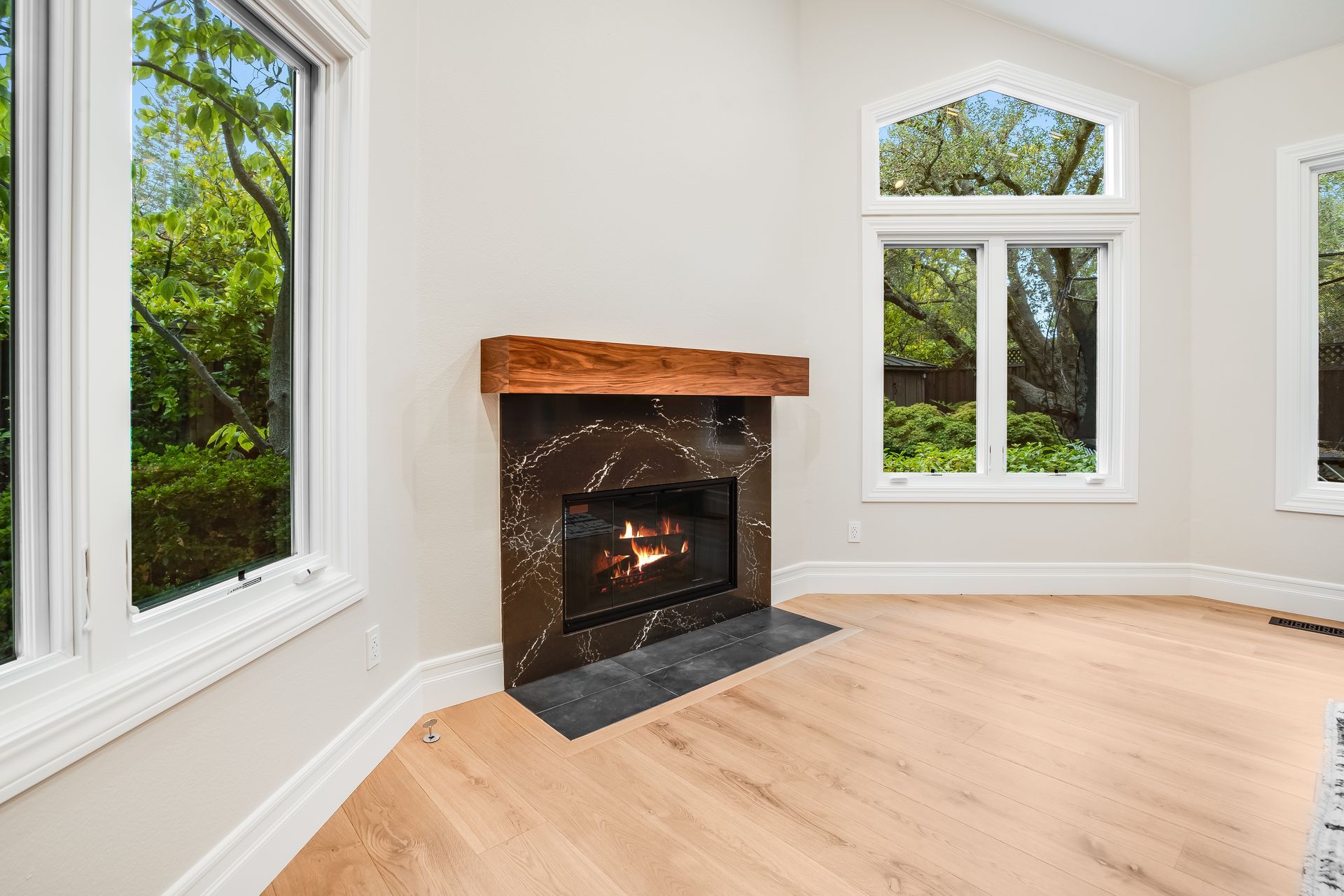 Bright living room with white walls, large windows, hardwood floors, and a fireplace with a dark stone hearth.