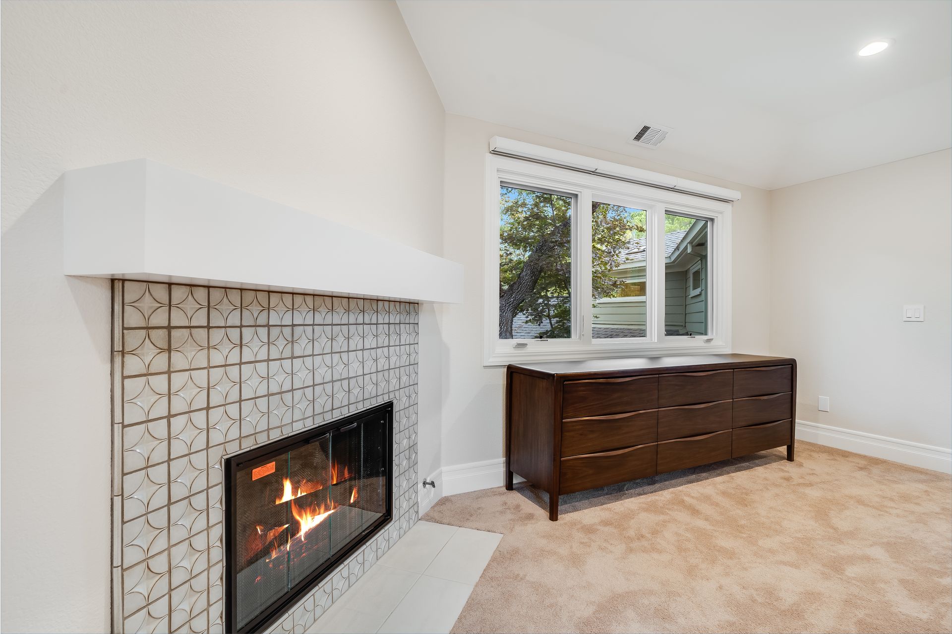 Bright living room with tiled fireplace, white walls, and dark wood dresser by windows
