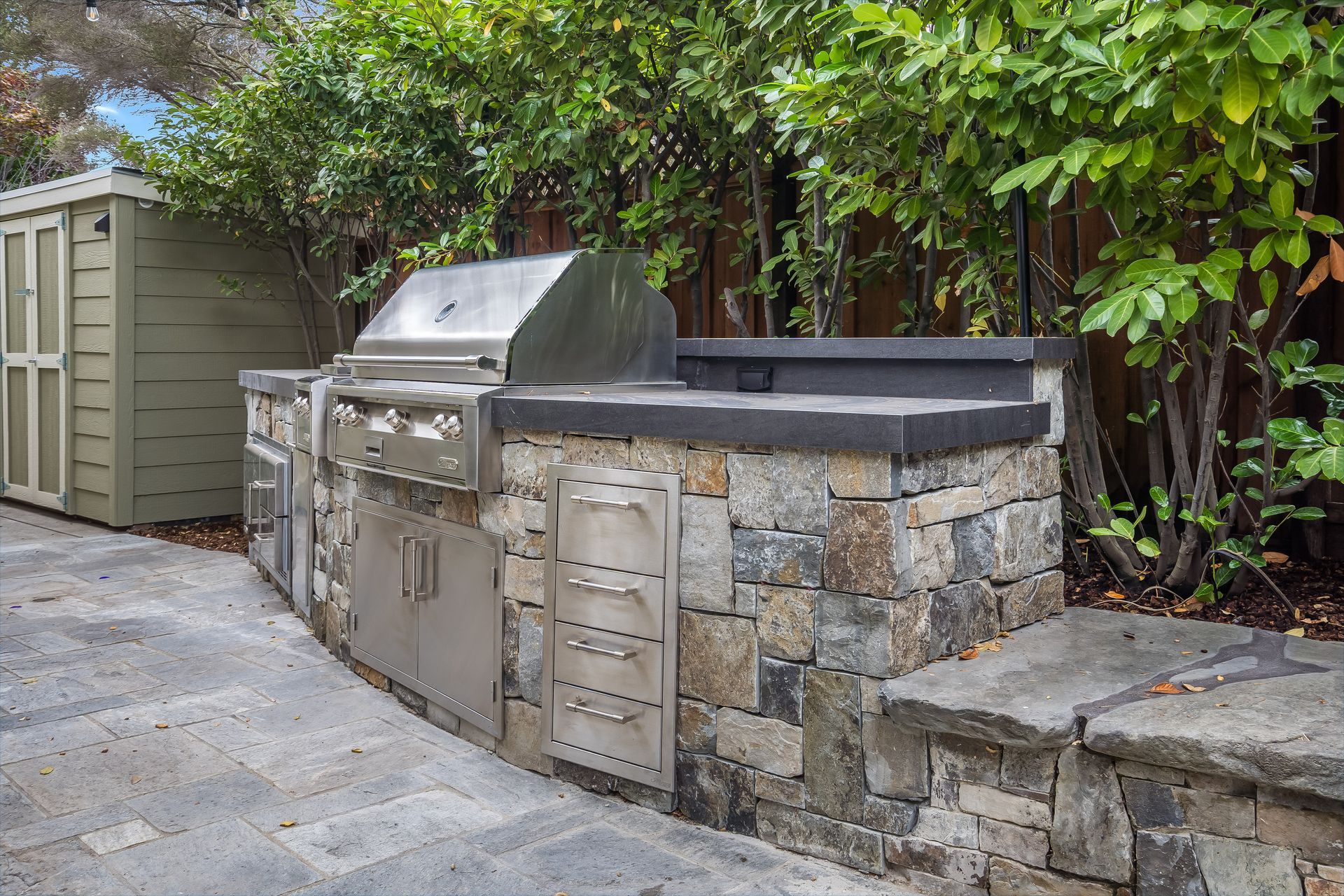 Outdoor stone barbecue grill with a metal lid beside a green shed and hedge-lined wall