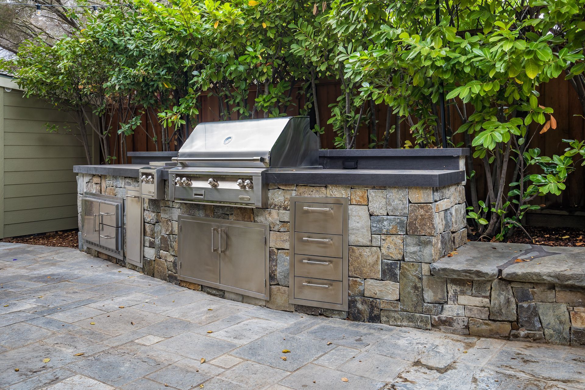 Outdoor stone patio with built-in stainless steel grill and cabinets under green shrubs