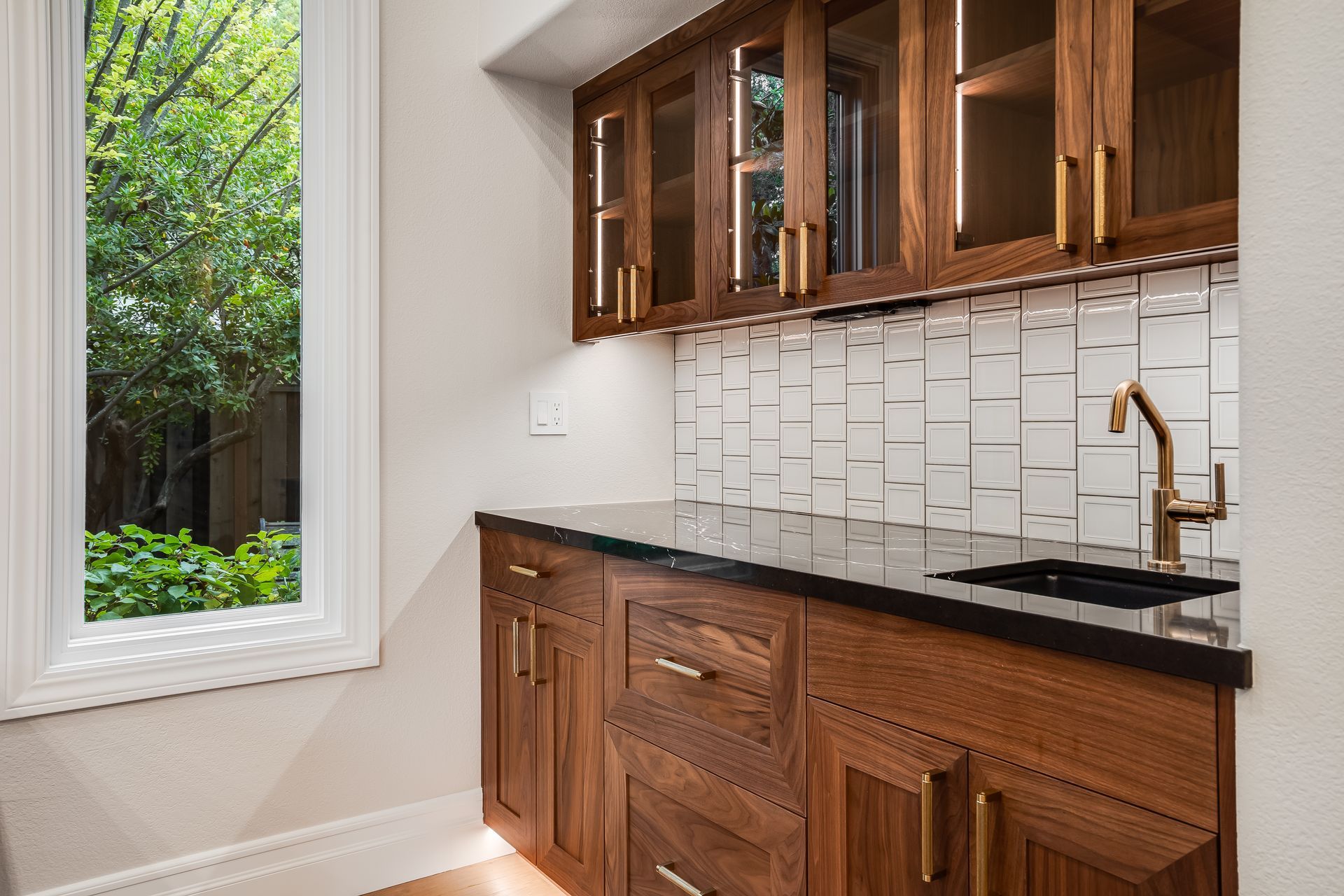 Wooden kitchen cabinets, black countertop, tiled backsplash, and a window overlooking greenery