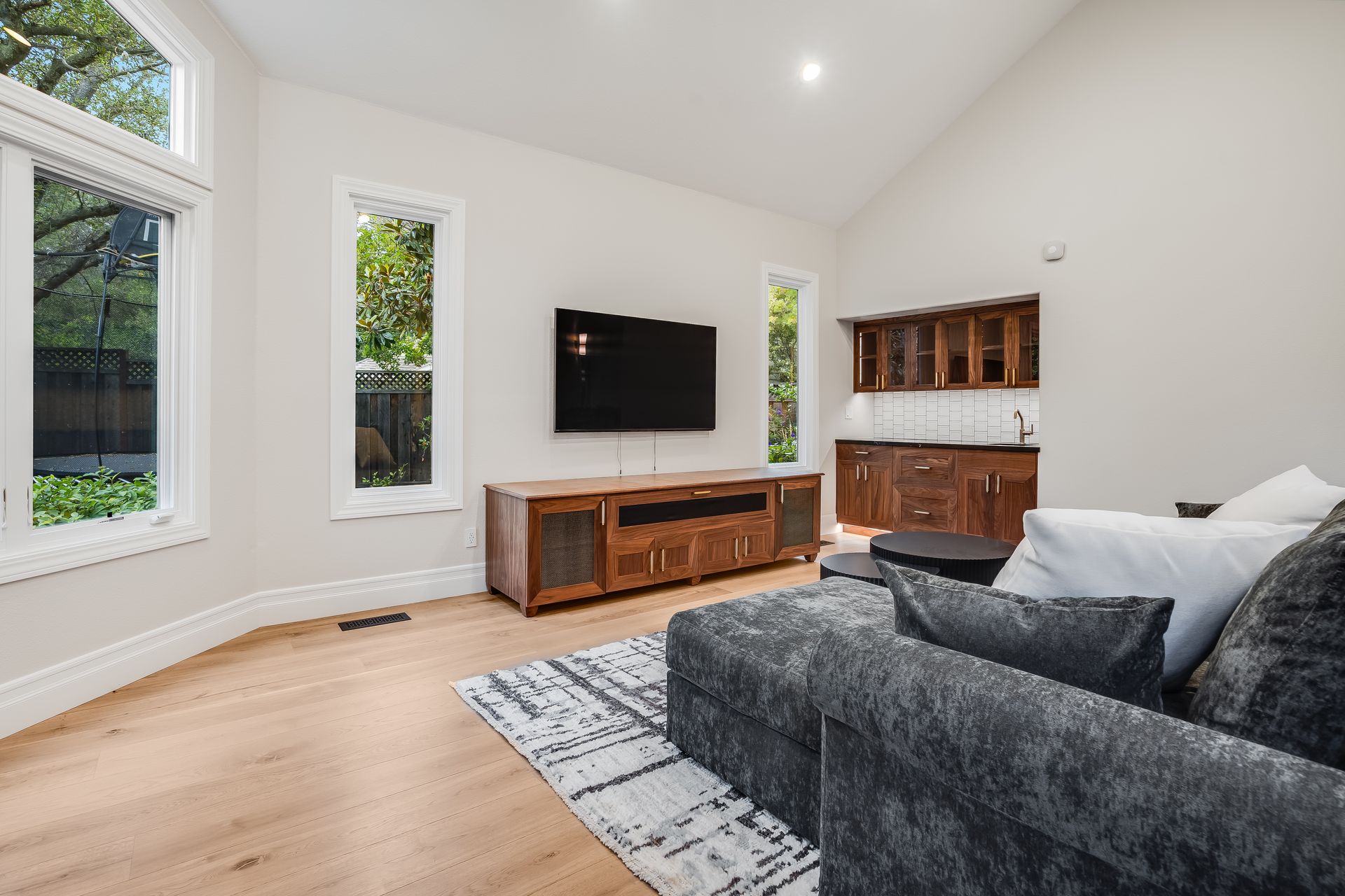 Bright living room with large windows, wall-mounted TV, wood media console, and gray sofa on a patterned rug