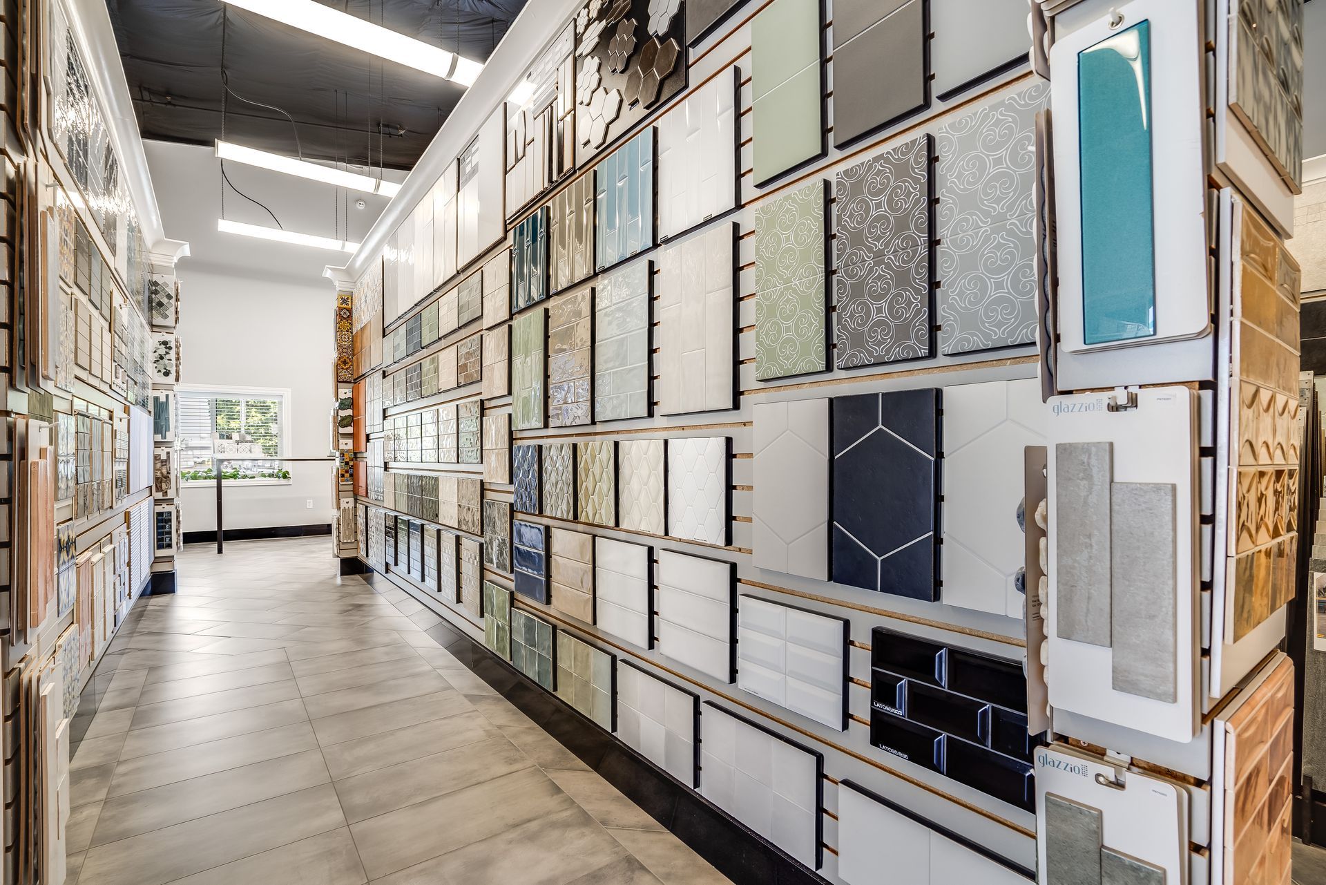 A tile showroom aisle with wall-mounted samples in many colors and patterns under bright lights.