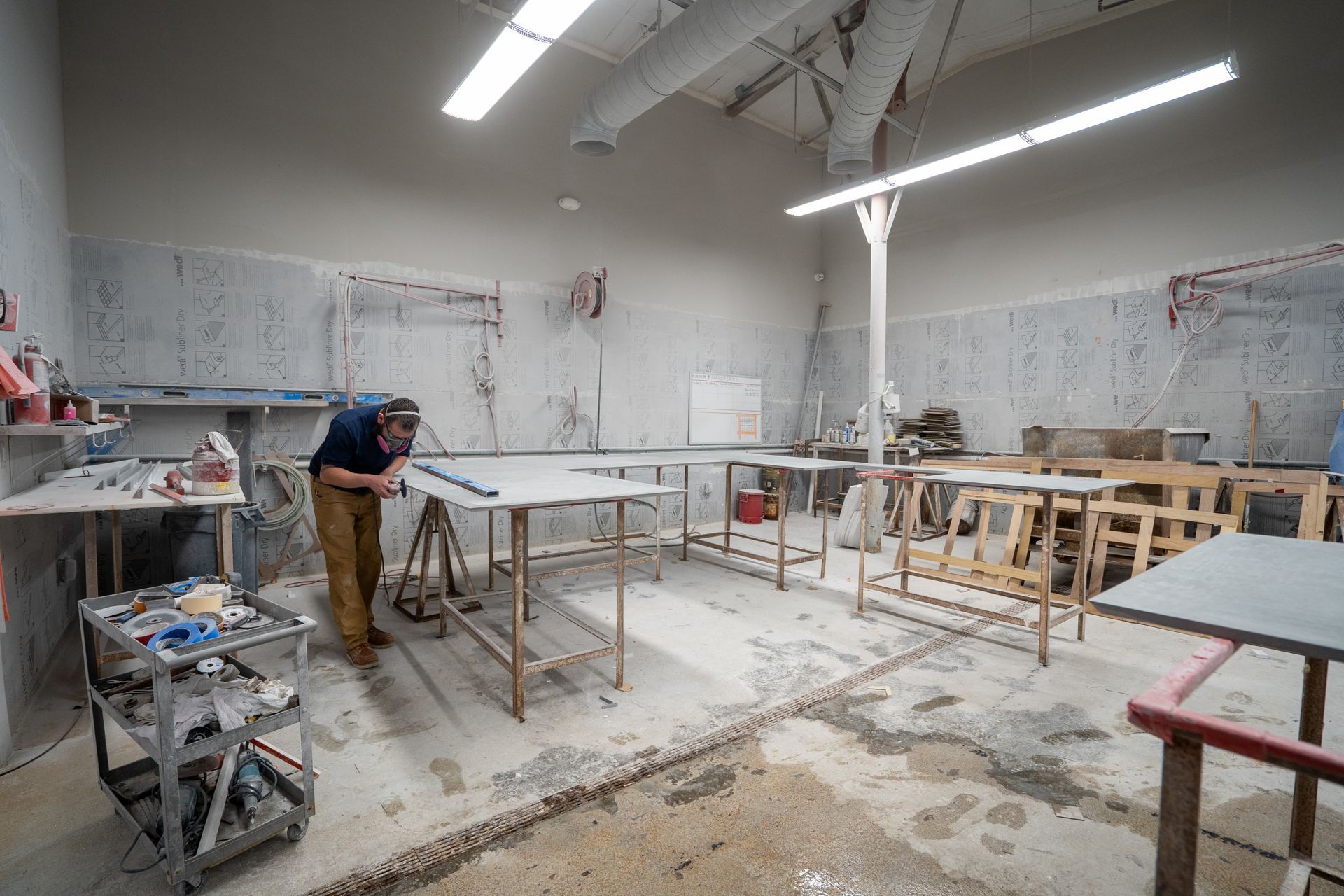 Person sanding a large table in a bright workshop with tools, shelves, and unfinished furniture.