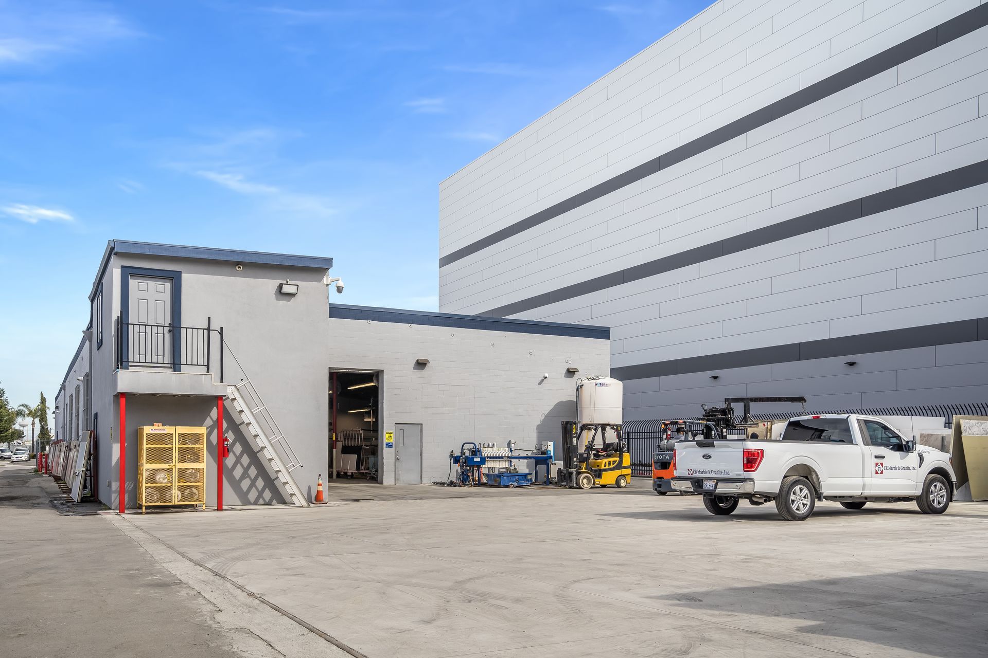 Outdoor industrial lot beside a gray warehouse, with parked pickup trucks and a small stairway building.